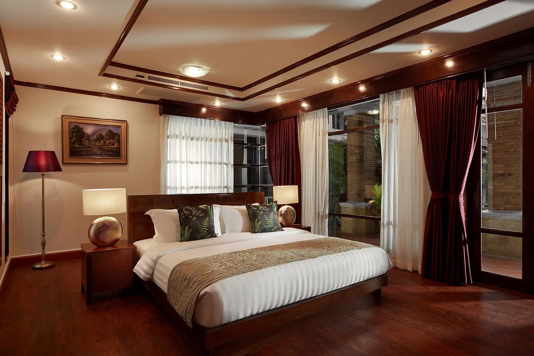 Luxurious villa bedroom with a large bed, nightstands with lamps, a framed landscape painting, dark red and white curtains, hardwood floors, and a view of a balcony with patio furniture. Best Phuket