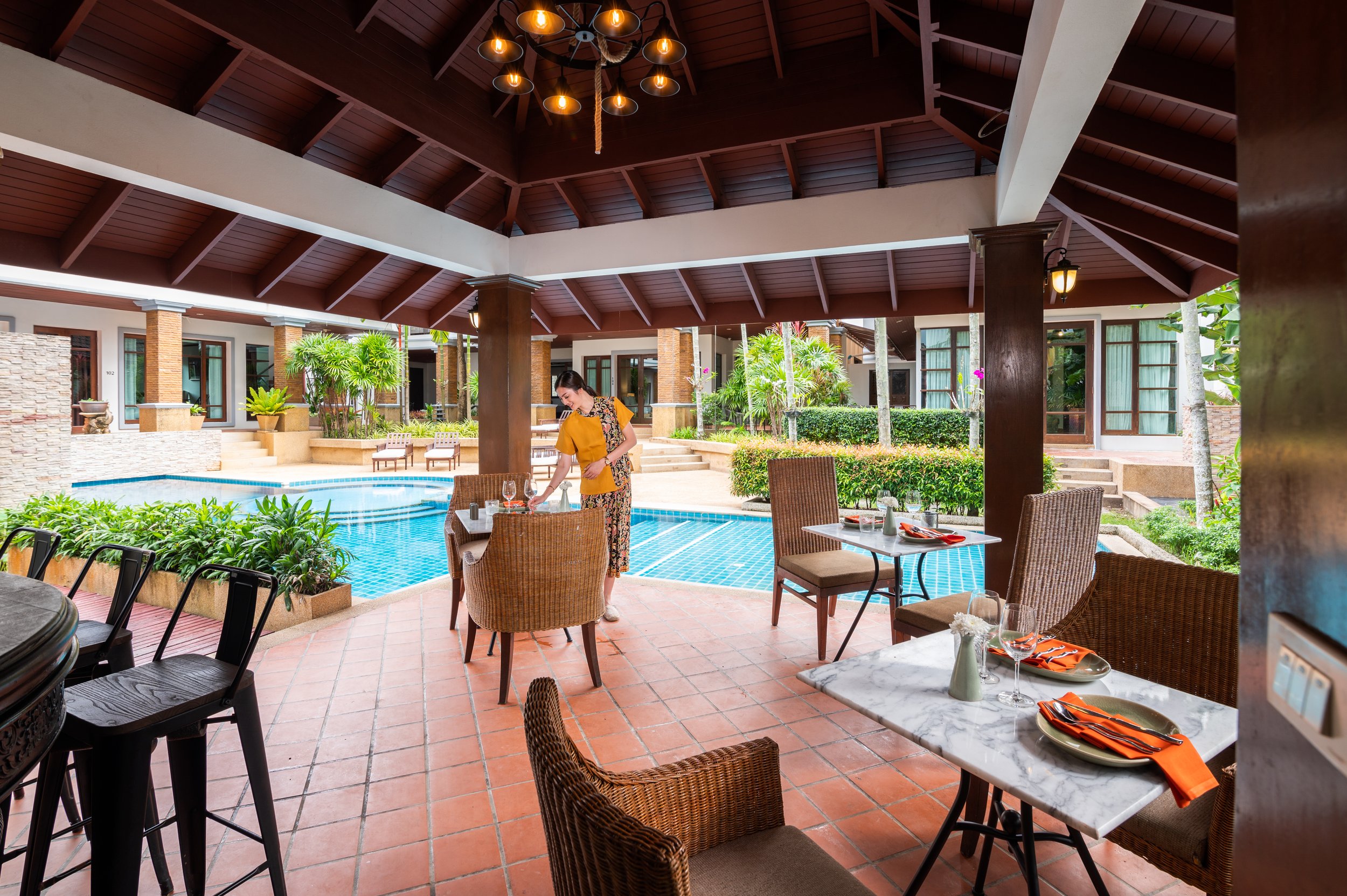 A woman setting a dining table in an outdoor restaurant area next to a swimming pool, with lush greenery and hotel rooms in the background.
