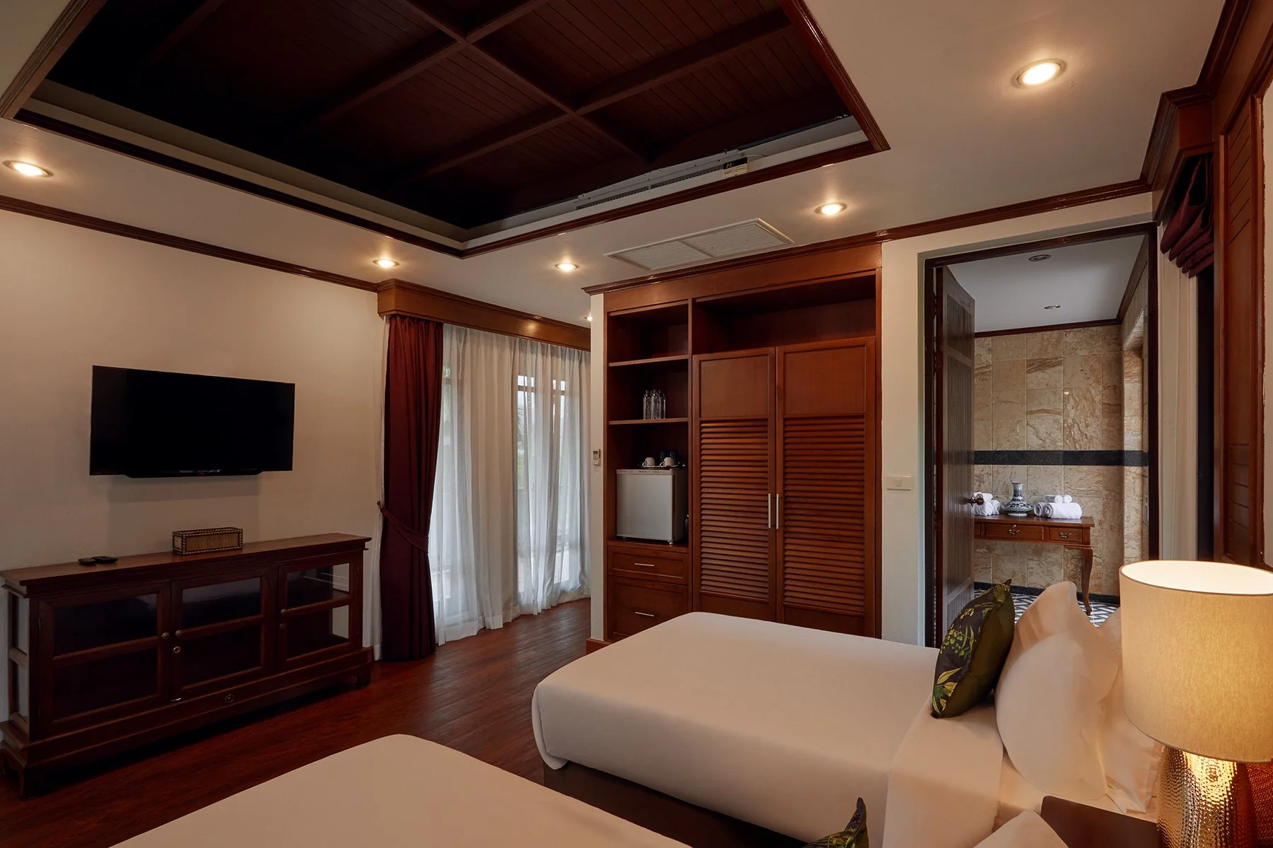 Villa room with two beds, a TV on a wooden stand, wooden wardrobe with a mini-fridge and glass bottles, window with curtains, and an en suite bathroom with a vanity and towel rack.