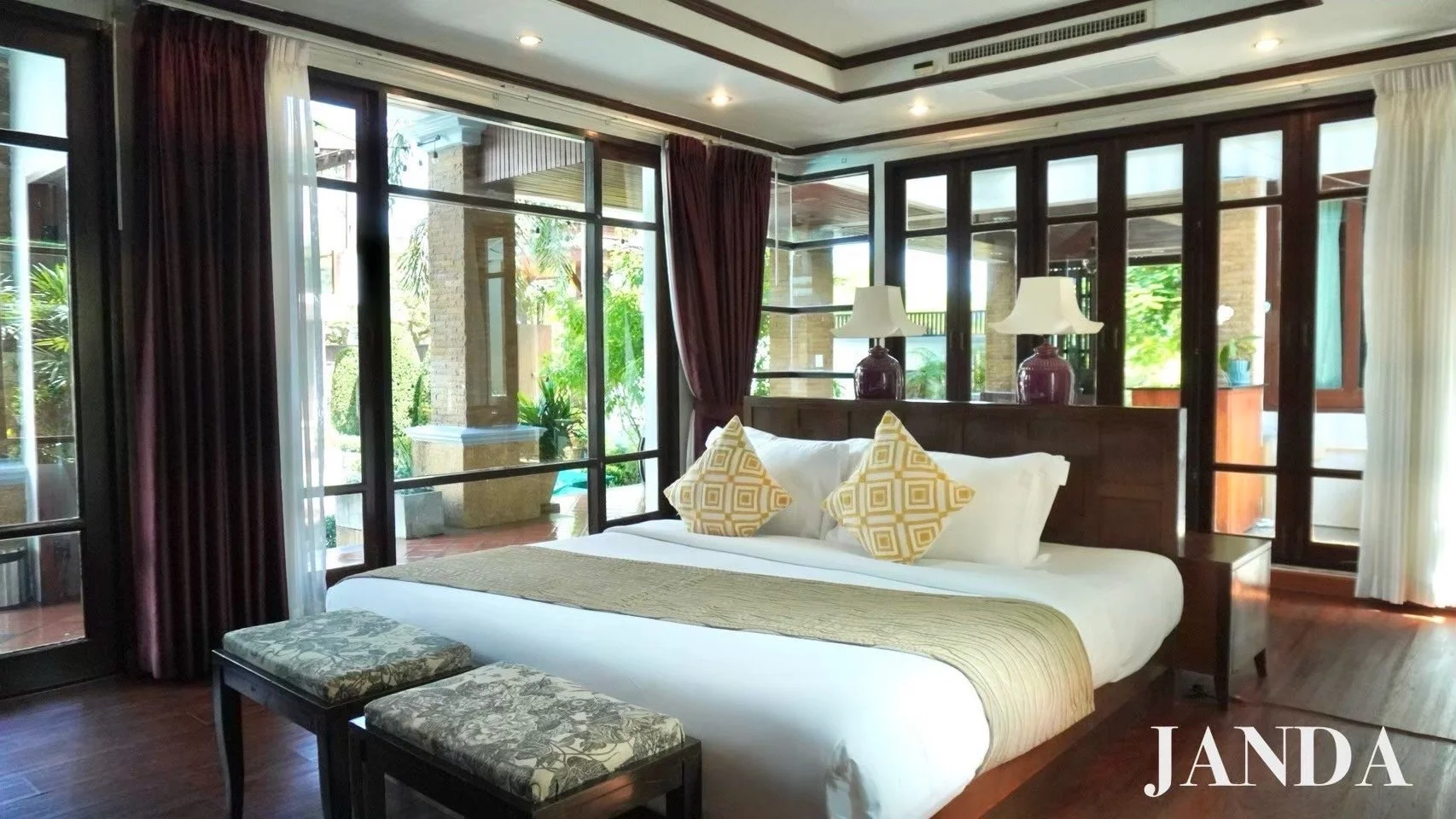 A luxurious bedroom with a large bed, decorative pillows, lamps, large windows with curtains, and outdoor greenery visible through the glass. The room has wooden floors and furniture.
