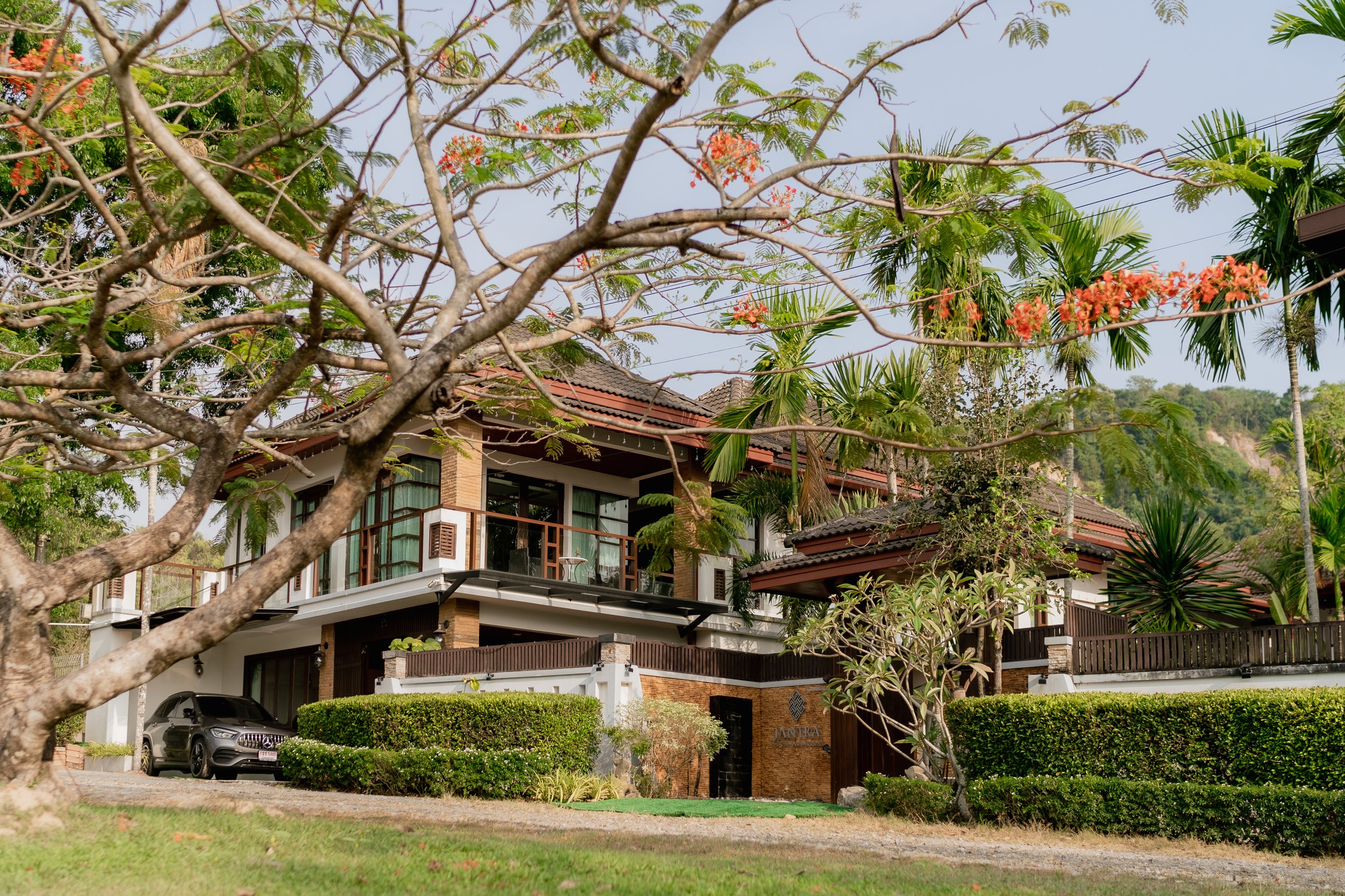 A modern two-story house with large windows, a balcony, and a garage partially visible behind landscaped bushes. The house is surrounded by lush tropical trees and plants, with one prominent tree in the foreground that has sparse branches and small o