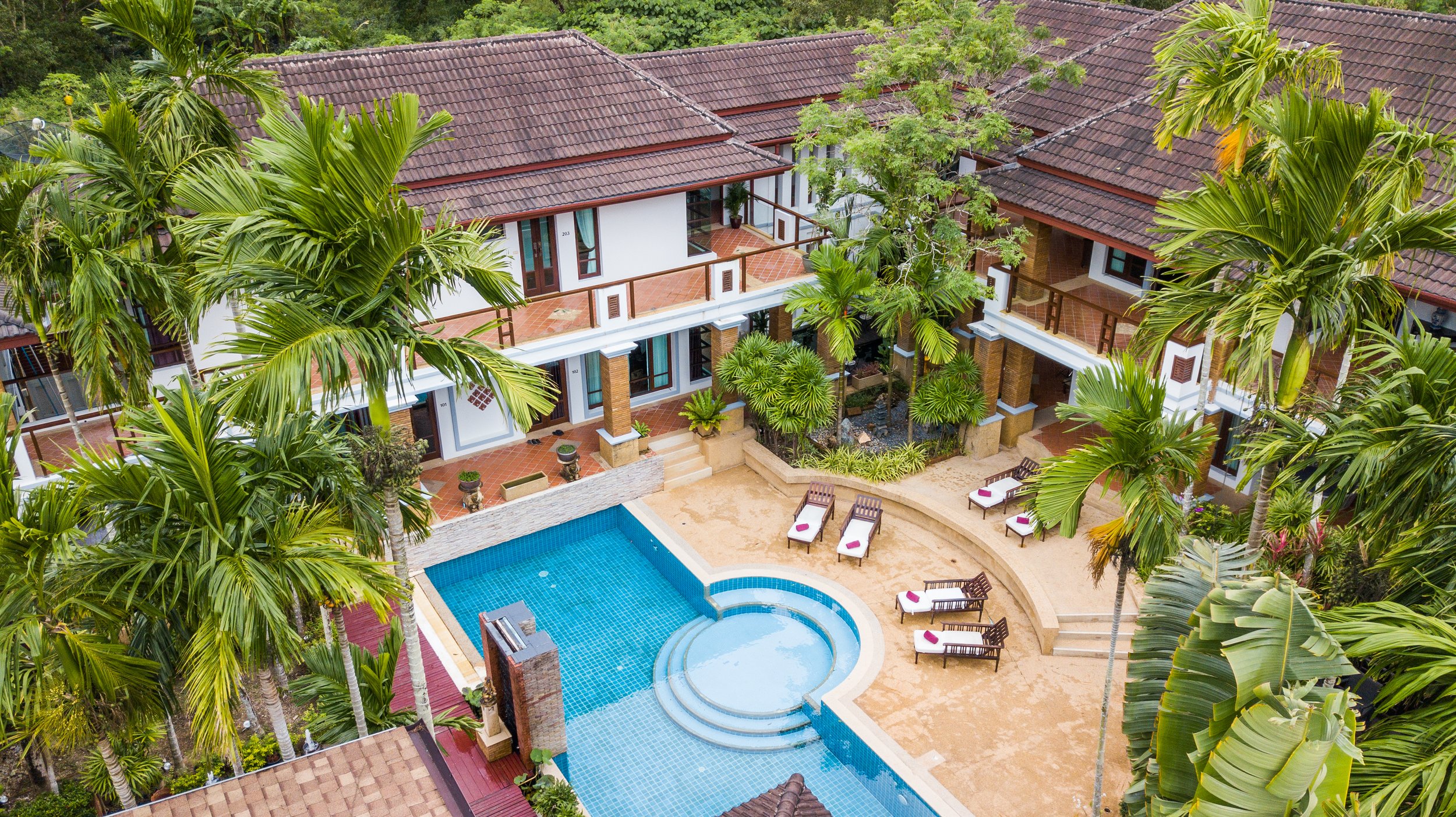 Aerial view of a tropical resort with a swimming pool surrounded by palm trees, lounge chairs, and a two-story building with balconies and tiled roofs. Large group villa Phuket