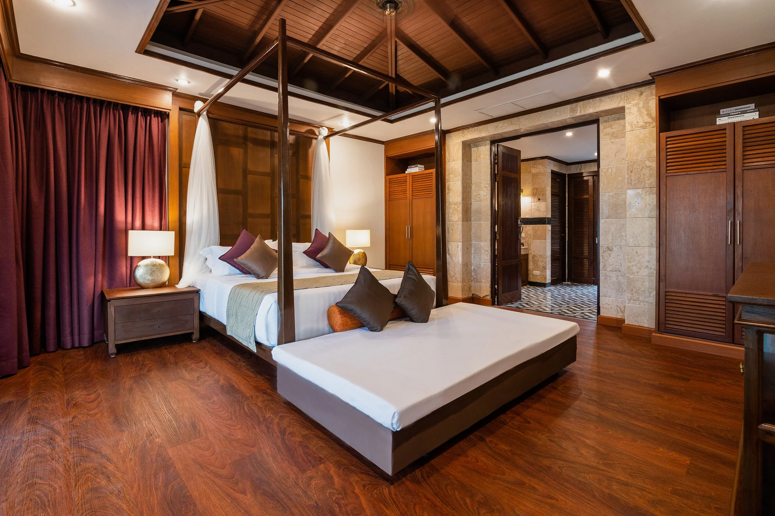 A luxurious hotel room with a canopy bed, wooden furniture, hardwood floors, and warm lighting. The bed has multiple decorative pillows, and there are lamps on the nightstands. The room opens to a corridor with tiled flooring.  Best high end villa