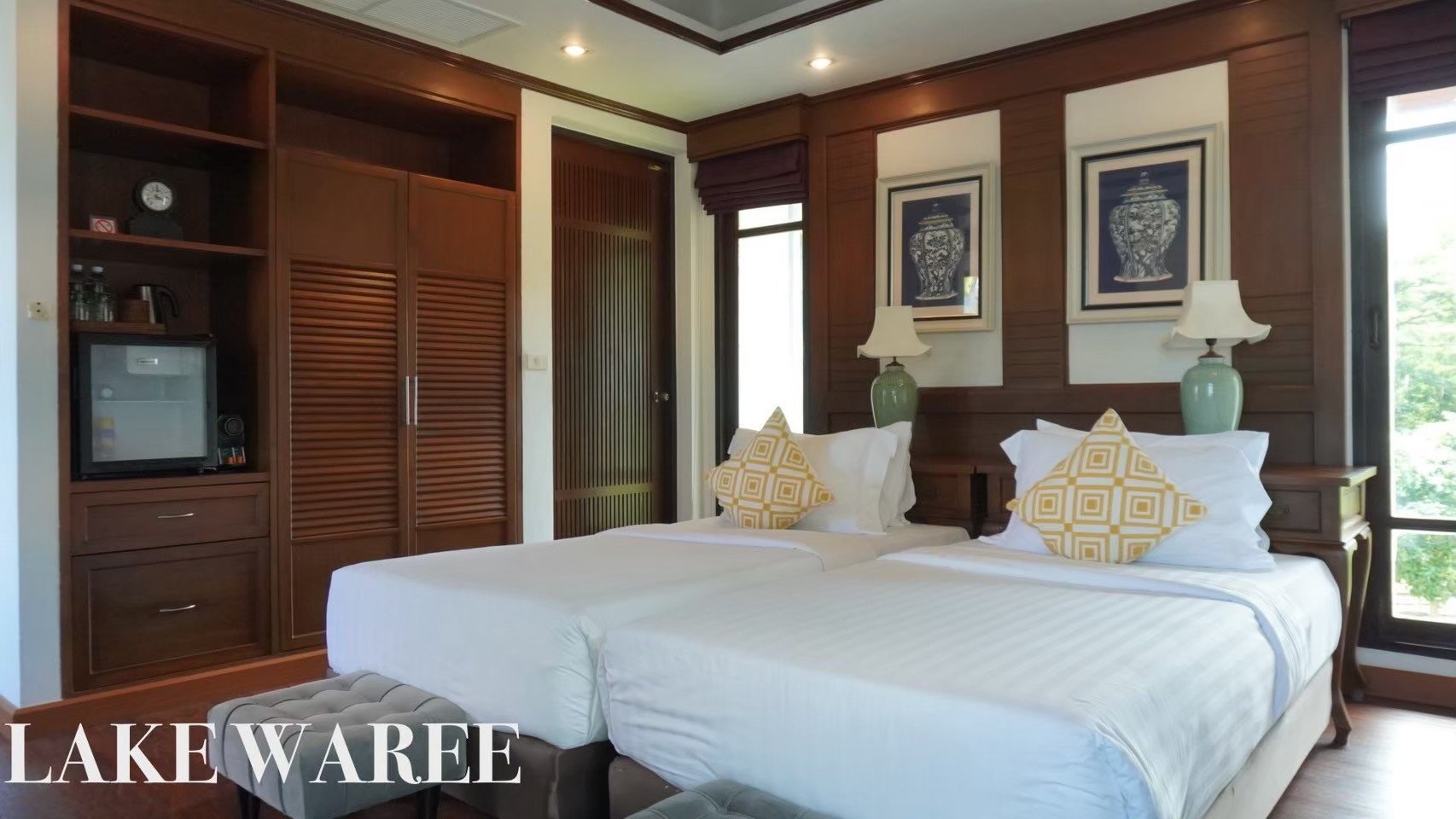 A bedroom with two twin beds pushed together, featuring white linens, yellow patterned pillows, and wooden headboards. Two green lamps are on bedside tables, and framed artwork hangs above the beds. The room has wooden paneling, a closet with louvere