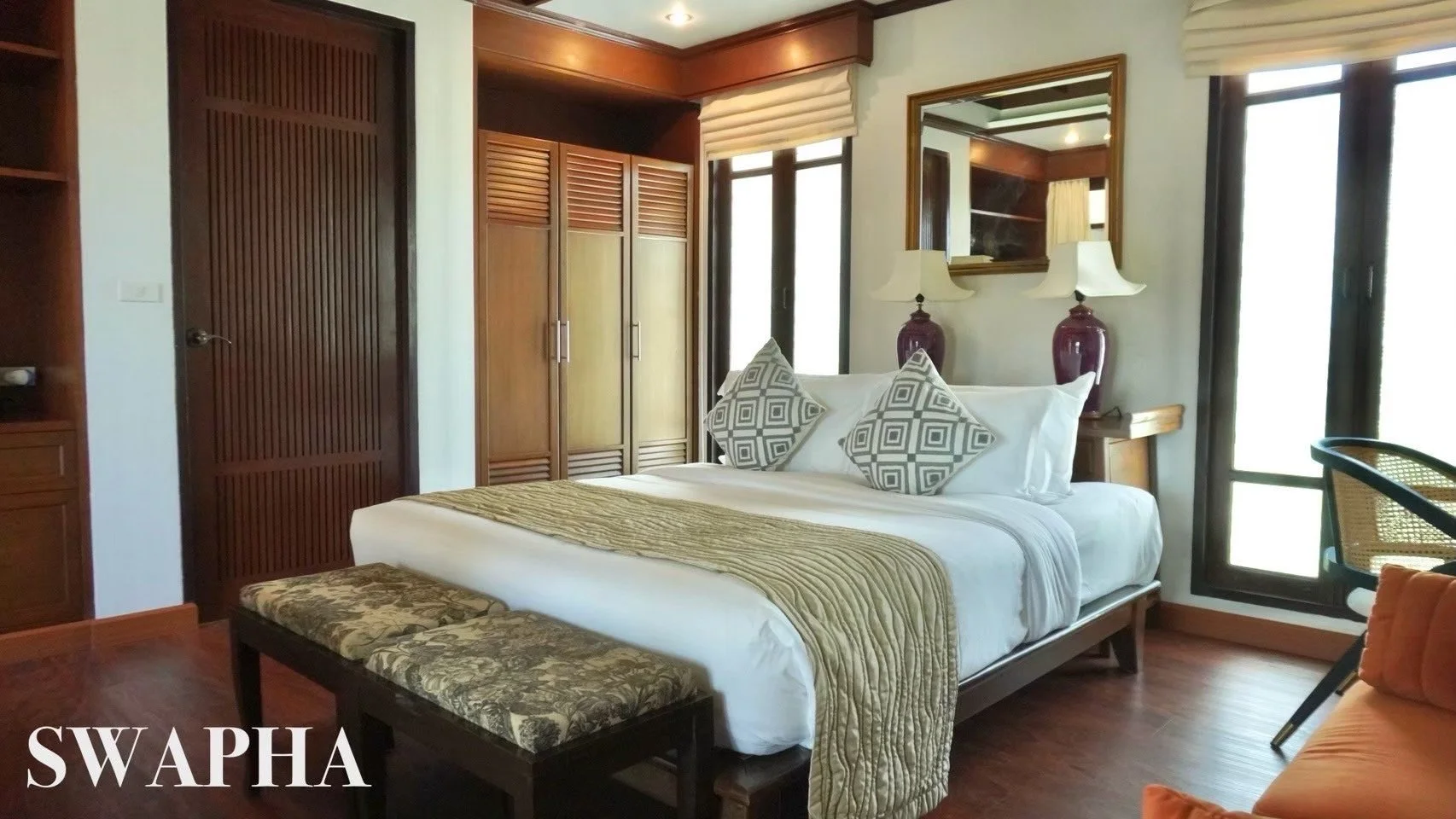 A bedroom with wooden furniture, a large white bed with patterned pillows and a decorative quilt, bedside lamps, a bench at the foot of the bed, and large windows with blinds.