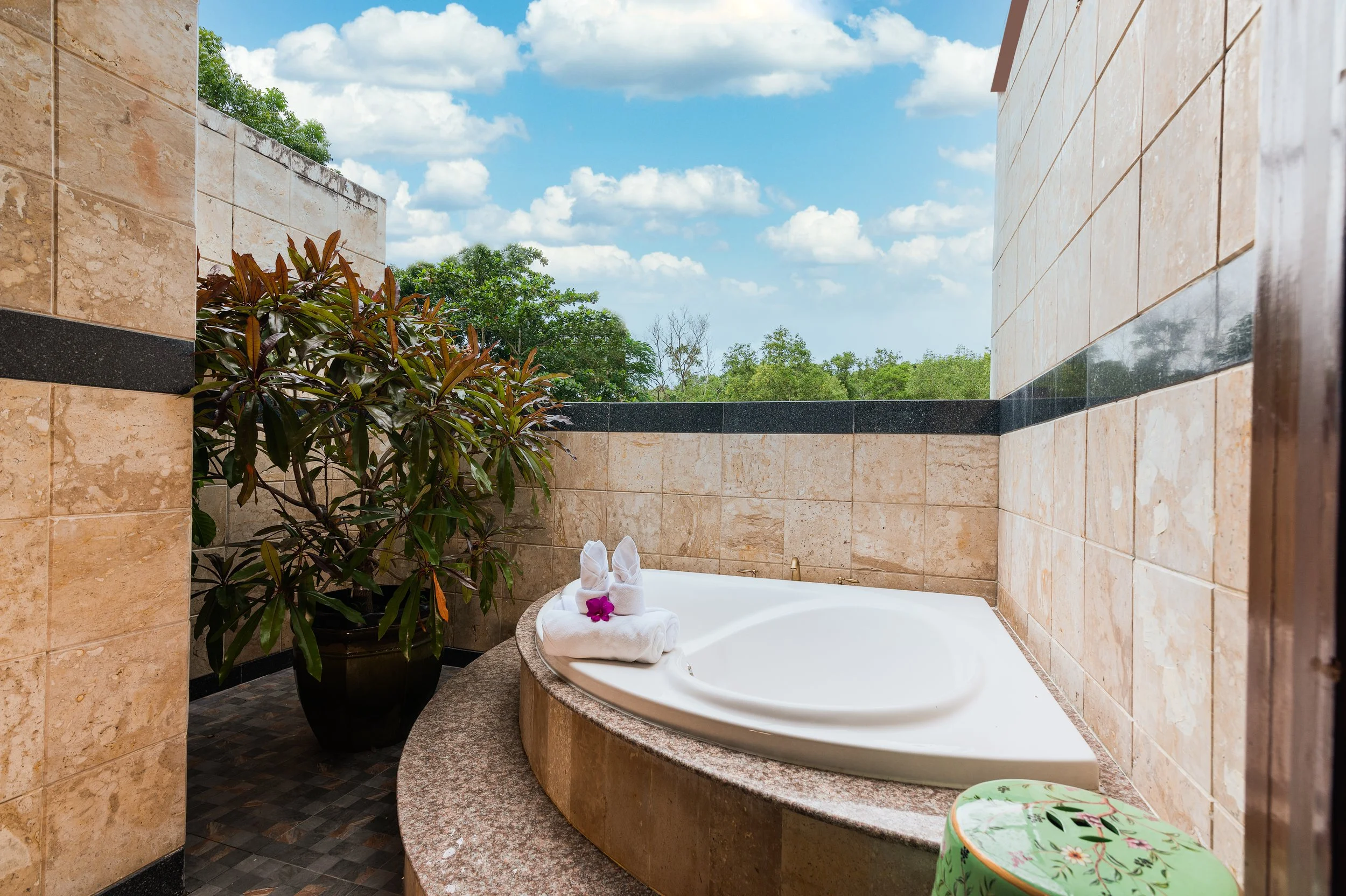 Outdoor bathtub in a private pool area with beige stone walls, a large potted plant, and a view of green trees and blue sky with clouds.