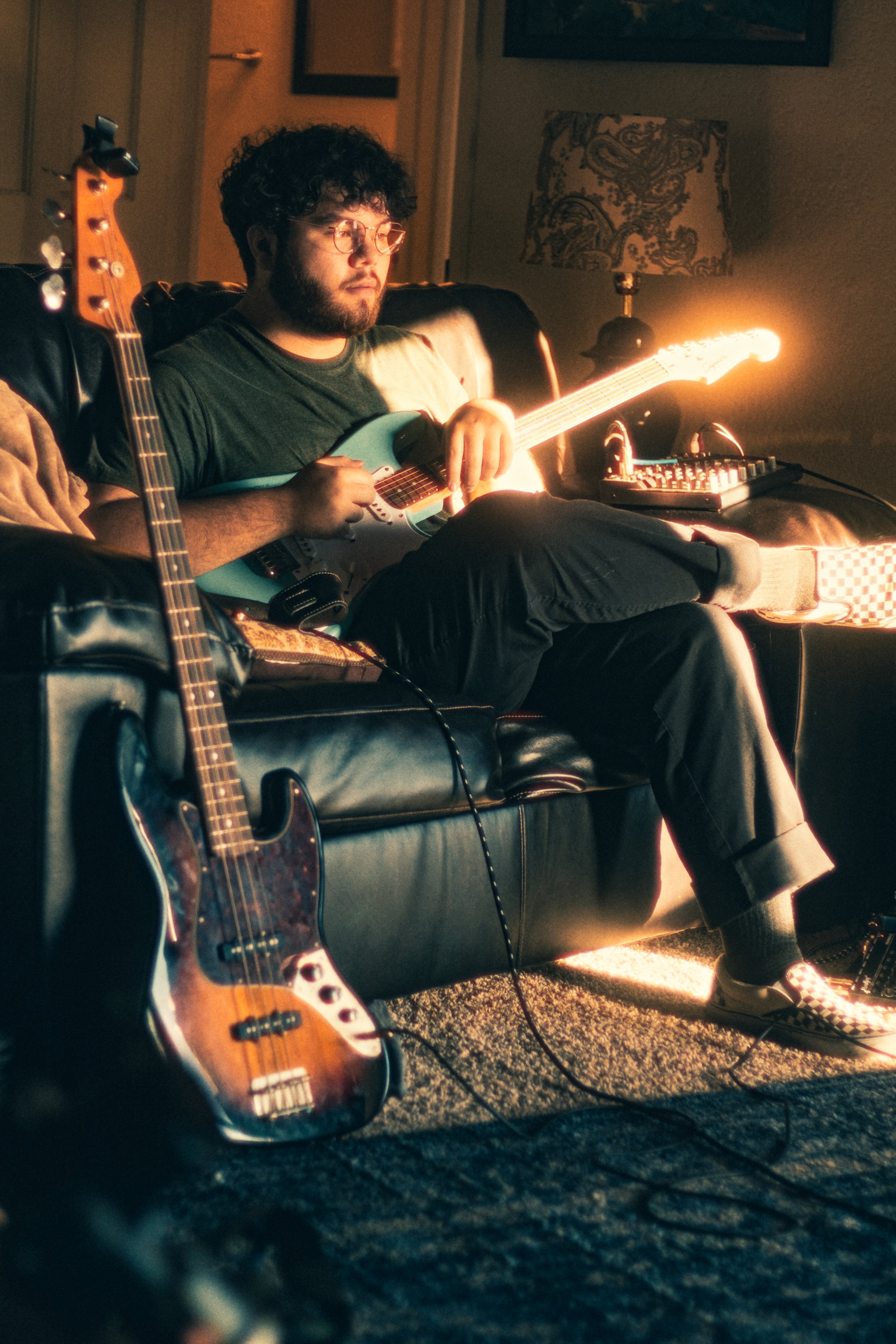 A young man with curly hair, glasses, and a beard sits on a black leather couch in a dimly lit room, playing an electric guitar. An electric bass guitar rests on the floor nearby, and a small music mixer is on the couch beside him. Warm, soft lighting illuminates the scene.