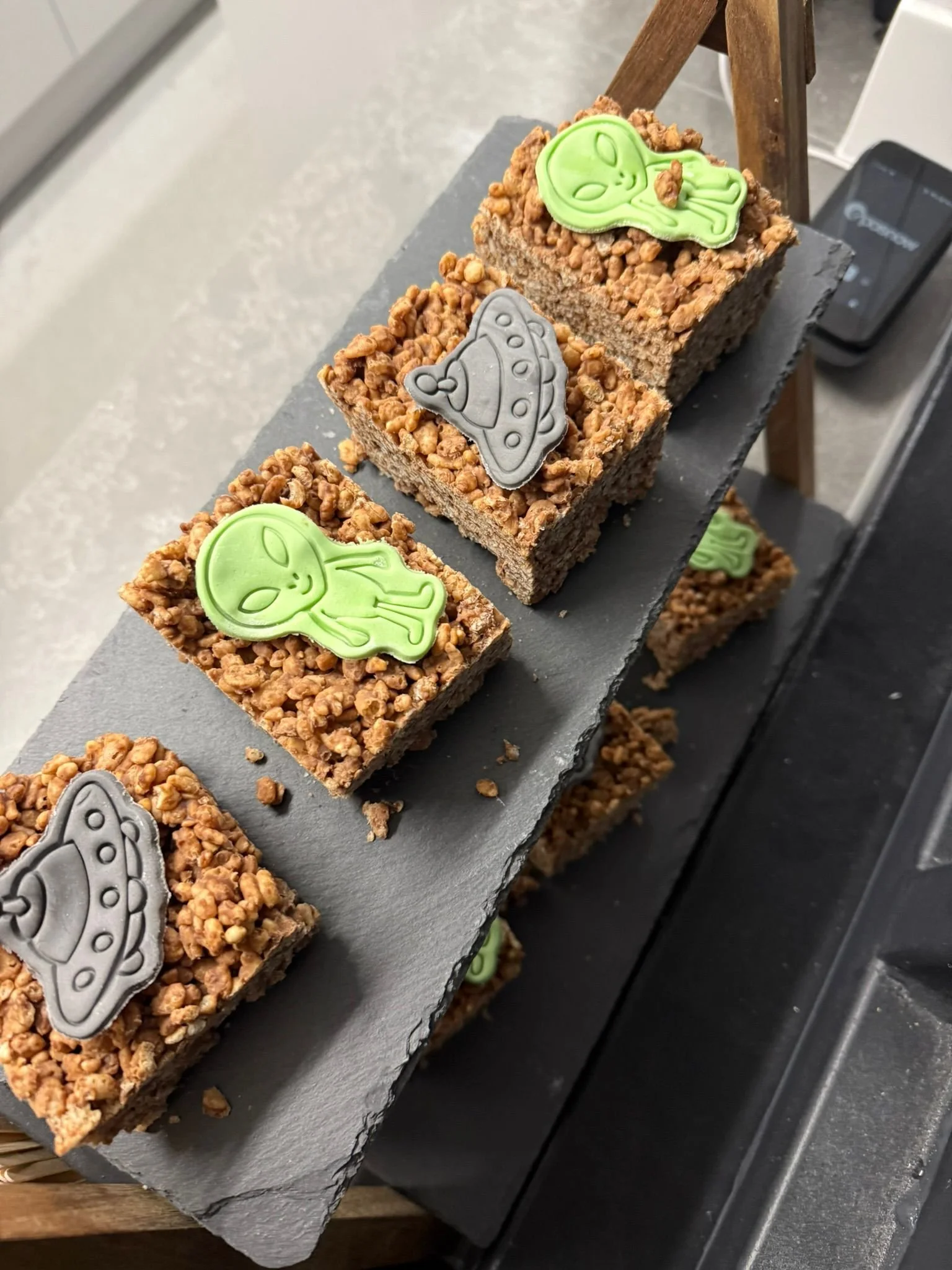 Multiple rectangular cereal bars topped with green and gray alien and spaceship shaped decorations, placed on a black slate stand.