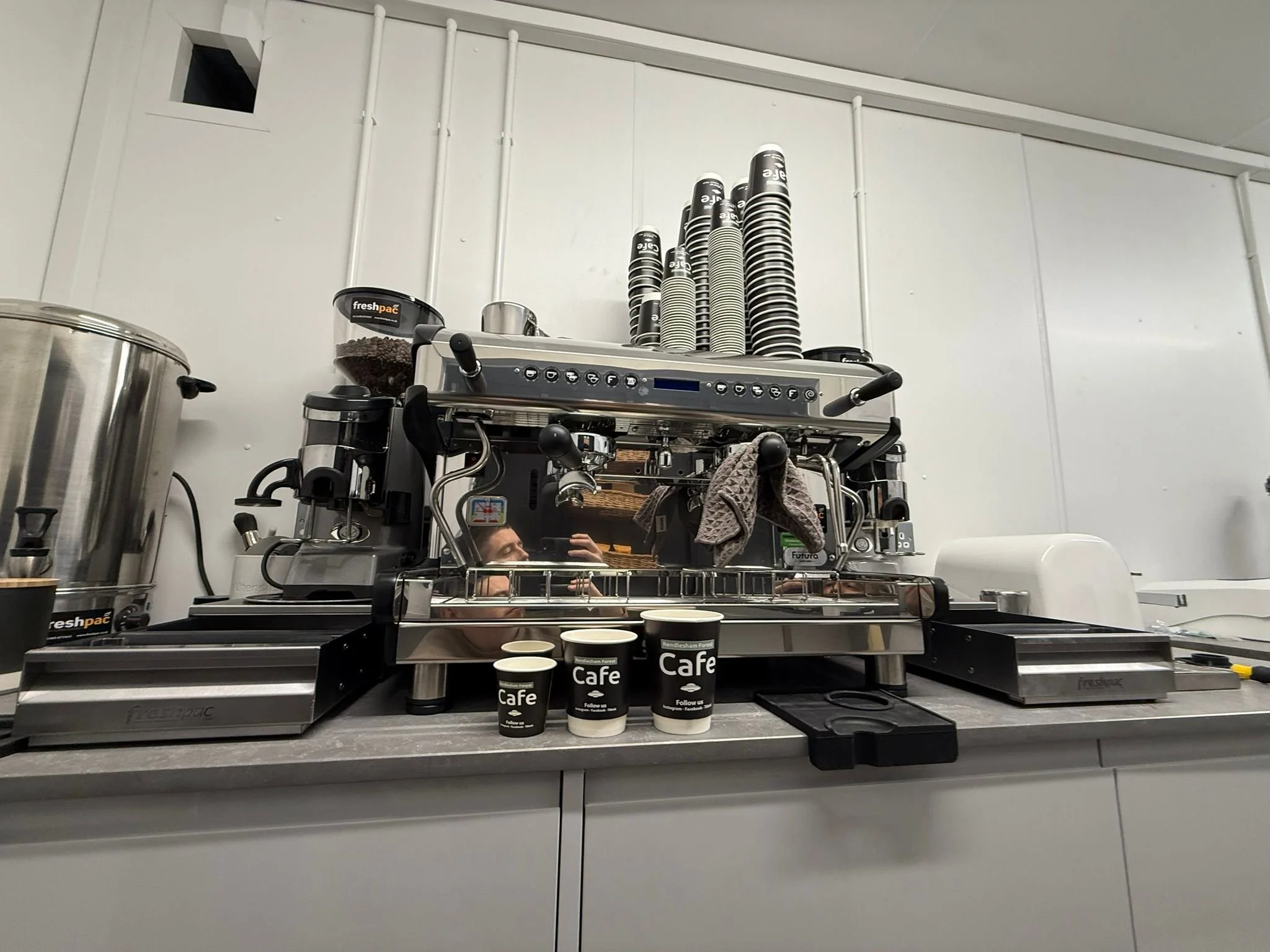 A commercial espresso machine with coffee cups on top, a counter with coffee cups labeled 'Cafe,' and a reflection of a person including hair and face in the shiny surface of the machine.