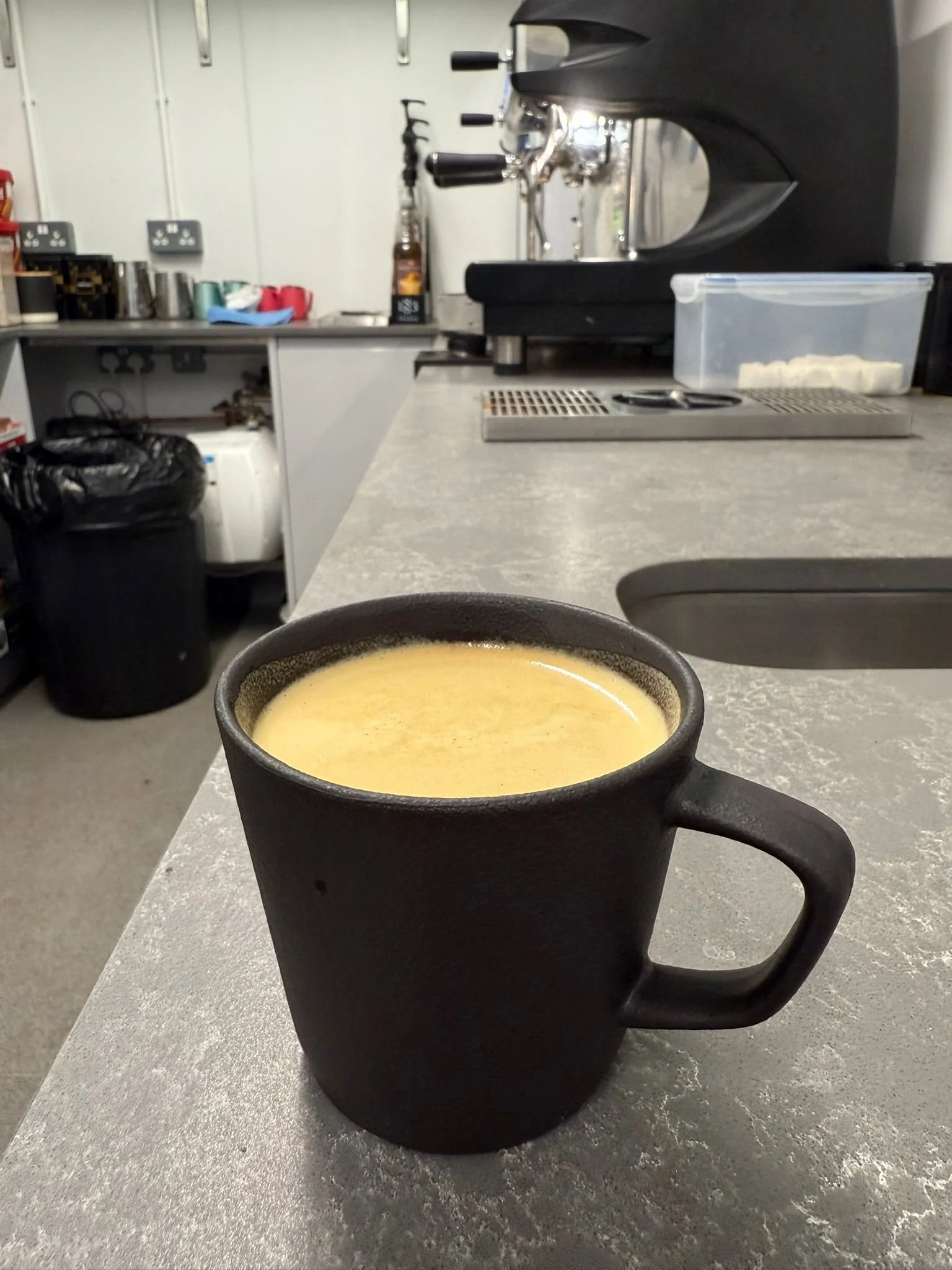 A black ceramic mug filled with a creamy yellow beverage, placed on a gray countertop in a kitchen or cafe setting.