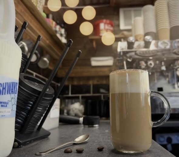 A tall glass mug filled with iced coffee topped with whipped cream, placed on a counter in a coffee shop. In front of the mug, there are a few coffee beans and a small spoon. The background shows shelves with cups, coffee stirrers, and other coffee shop supplies.
