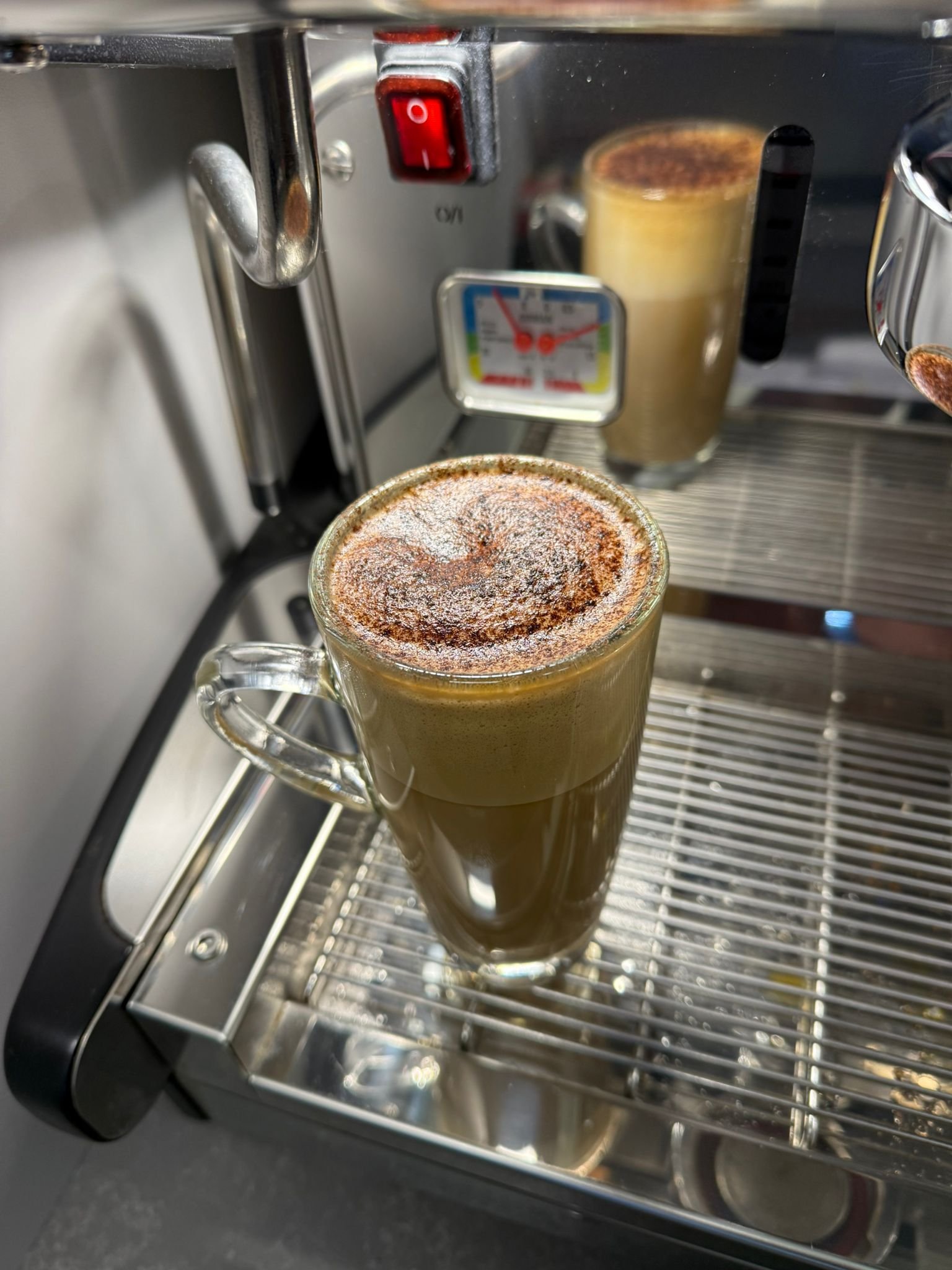 A glass mug of freshly brewed cappuccino with a dusting of cocoa powder on top, sitting on a metal drip tray of an espresso machine.
