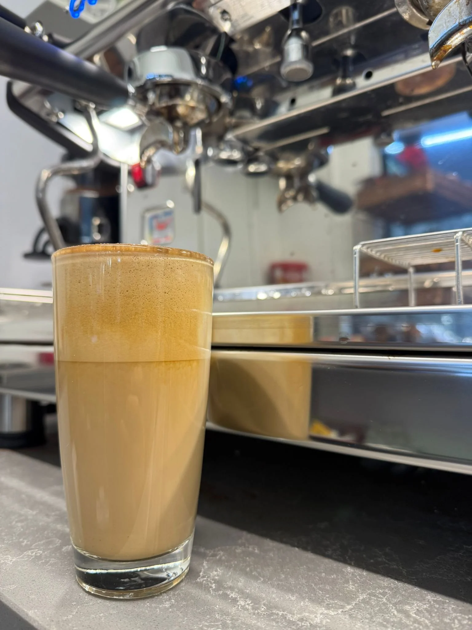 A close-up of a glass of frothy coffee or latte on a gray countertop with a commercial espresso machine in the background.