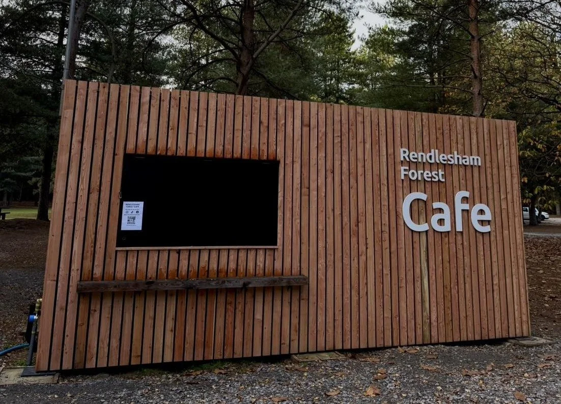 Wooden sign at Rendesham Forest Cafe with a mounted black screen and a QR code, surrounded by trees.