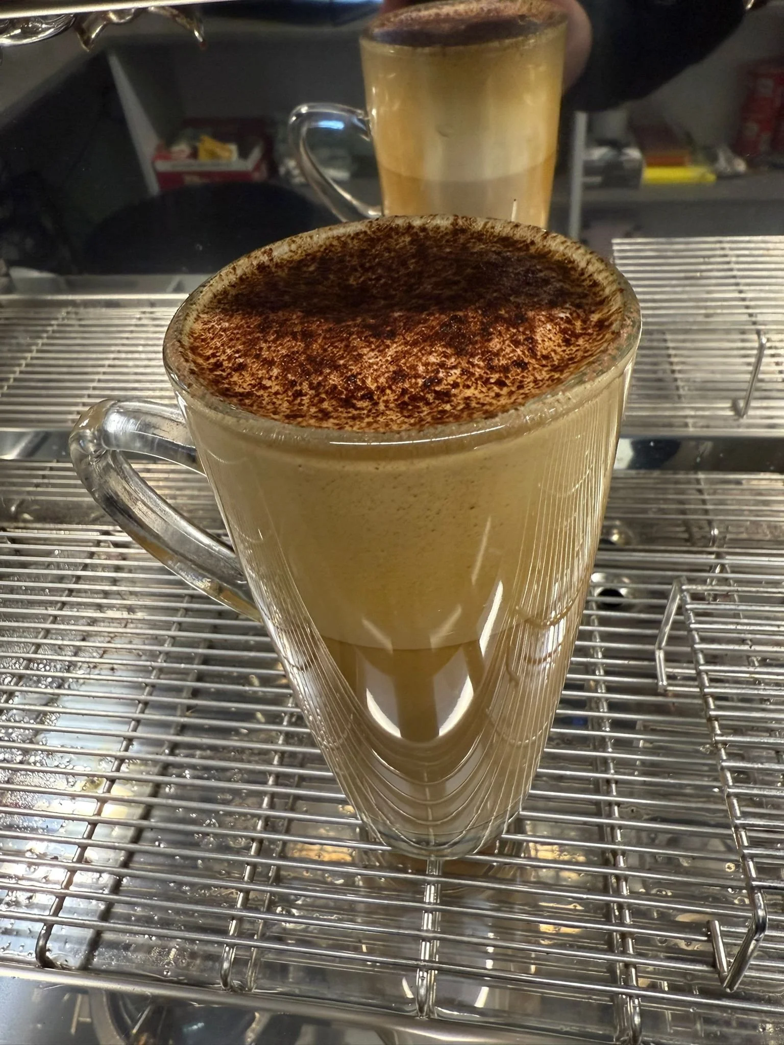 Two glass cups filled with topped with frothy, coffee-flavored beverages, possibly cappuccinos or lattes, sitting on a metal wire rack in a kitchen or café setting.