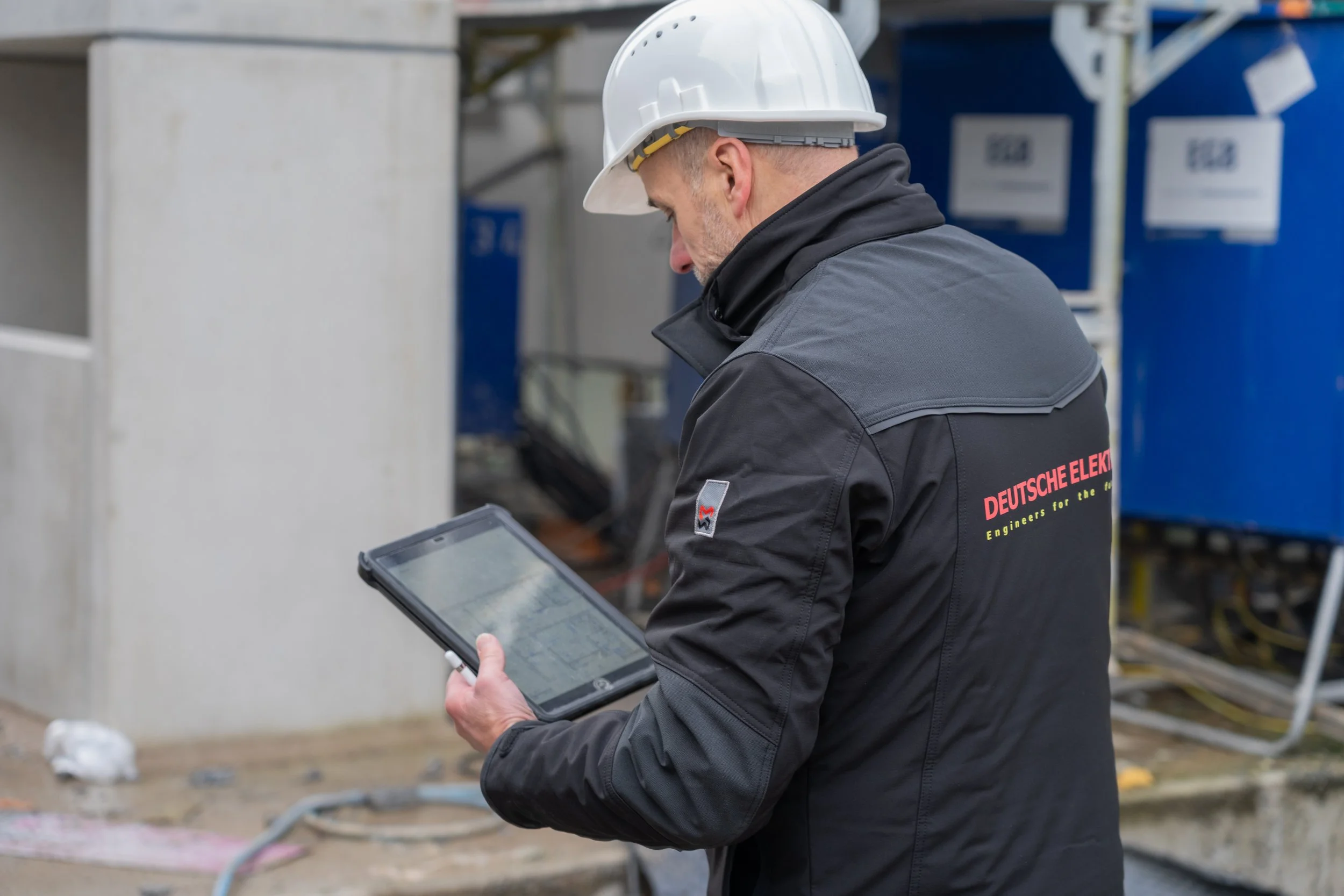 Ein Mann mit Schutzhelm und Arbeitsjacke, die mit "Deutsche Elek" beschrieben ist, benutzt ein Tablet bei einer Baustelle.