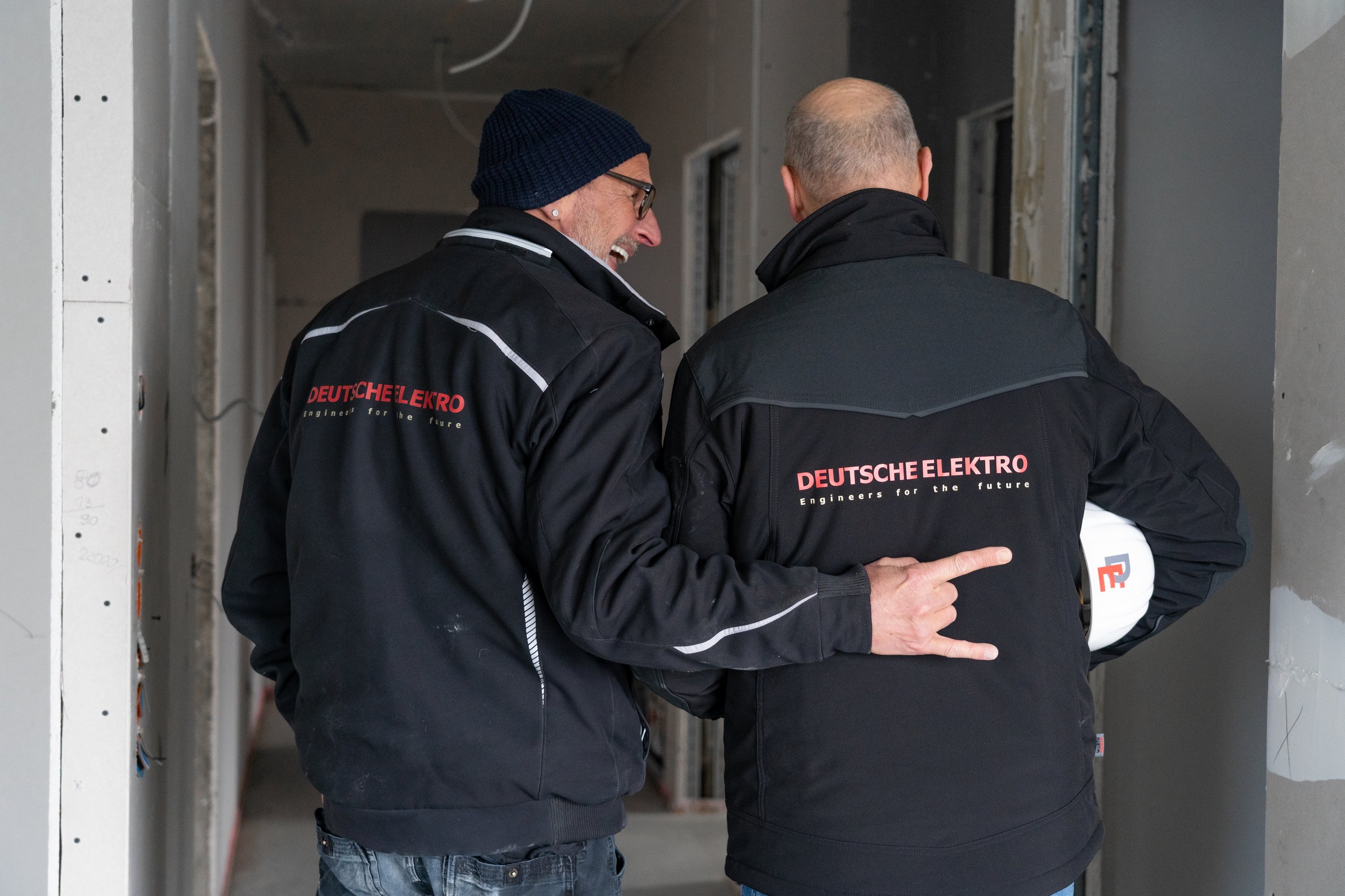 Zwei Männer in dunklen Jacken mit 'DEUTSCHE ELEKTRO' auf dem Rücken, im Gespräch in einem Baugerüst oder Rohbau.