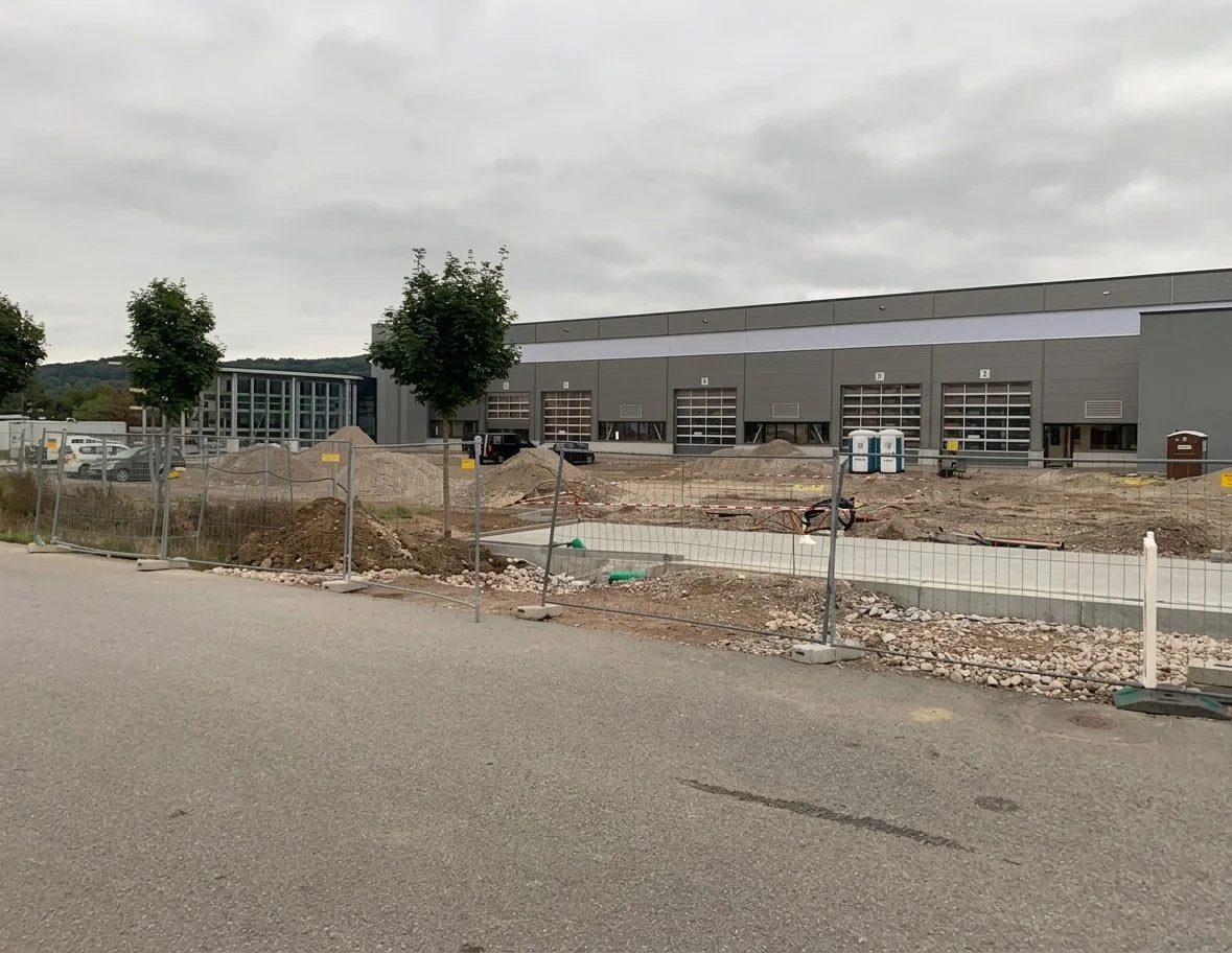 Bauarbeiten vor einem modernen Industriegebäude, mit Baggerloch, Bauschutt und Bauzäunen im Vordergrund.