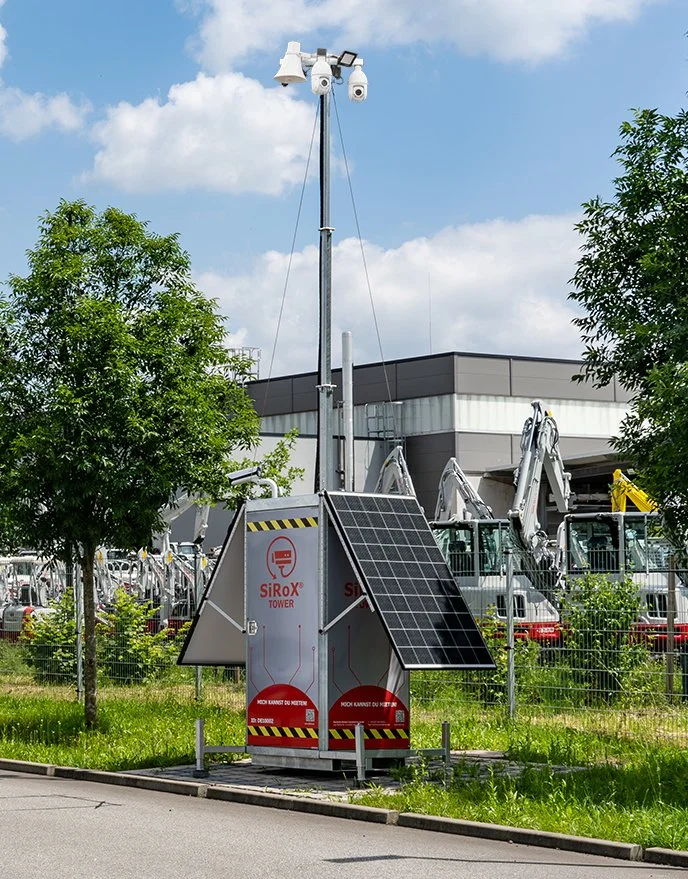Solarbetriebener Überwachungsturm mit Messgeräten und Solarpanel, umgeben von Bäumen und Gebäuden.