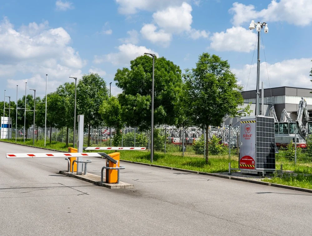 Ein geschlossener Parkplatz mit einen Schranken, einige Bäume, Laternen, und eine Sicherheits- oder Energiebox im Vordergrund, im Hintergrund eine Reihe von Baugeräten und einem modernen Gebäude.