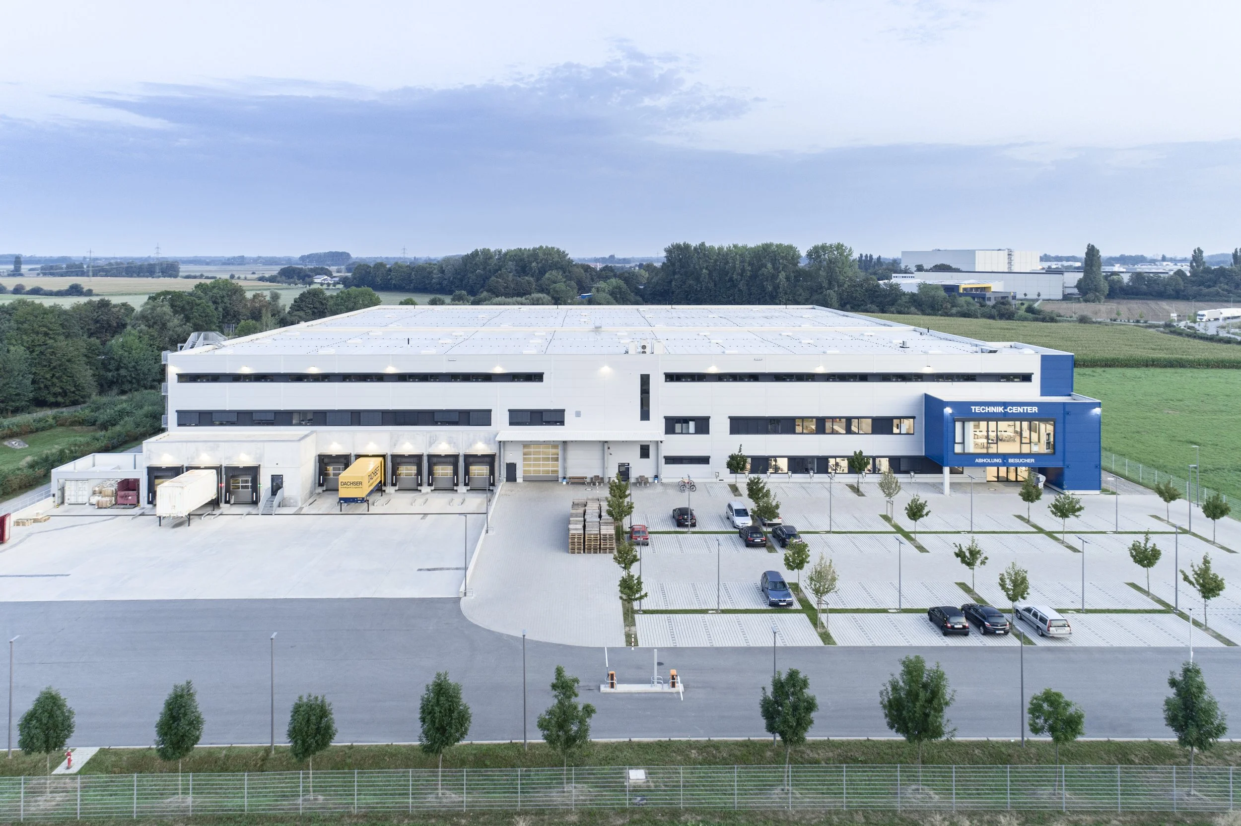 Luftaufnahme eines modernen Industrie- oder Logistikzentrums mit weißen Gebäuden, einem Parkplatz mit wenigen Autos und umgeben von grünen Feldern und Bäumen.