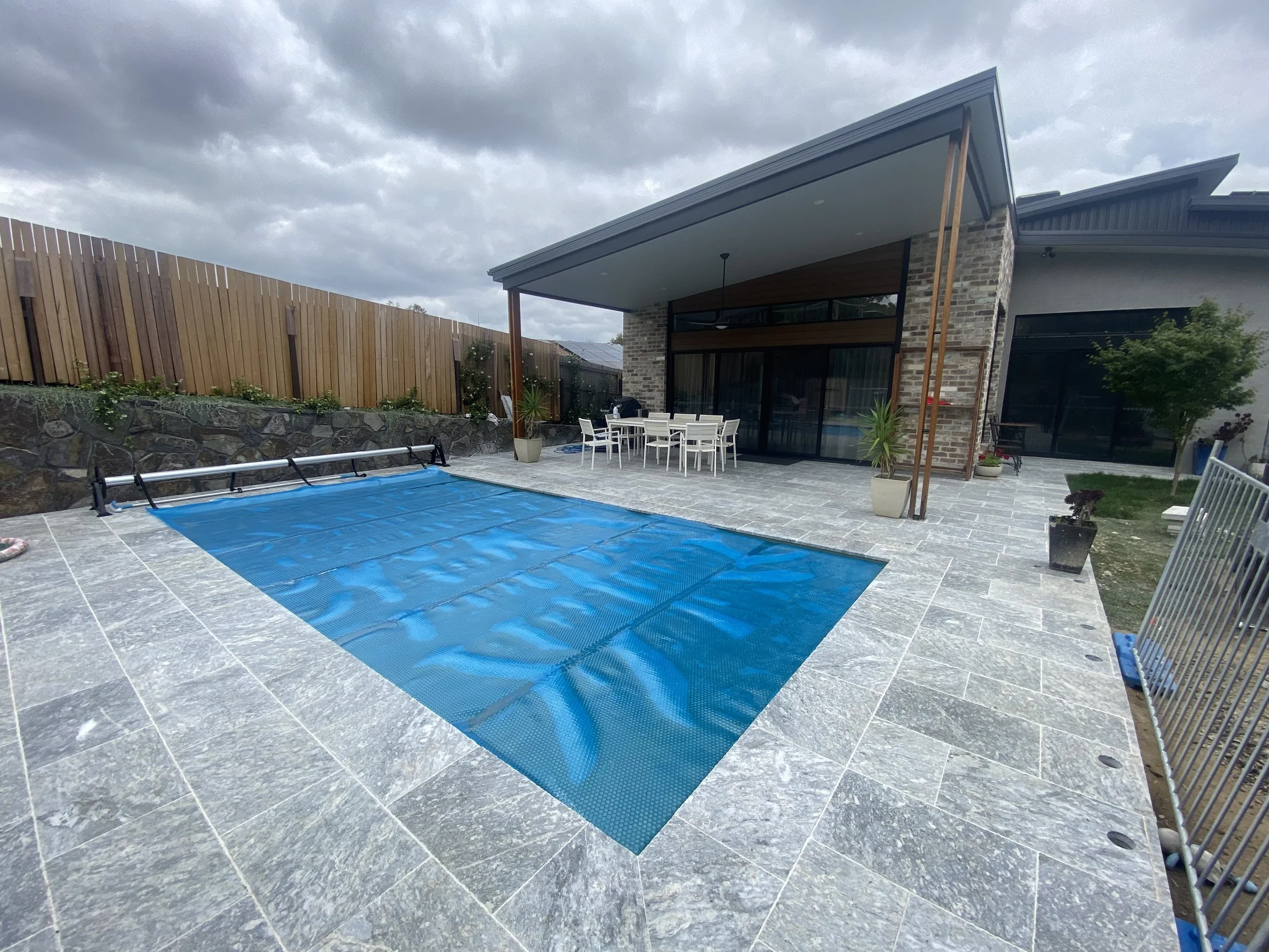 Backyard view showing a covered patio with dining table and chairs, a pool covered with a blue safety cover, potted plants, and a stone patio in front of a modern house with brick and wood accents, under a cloudy sky.