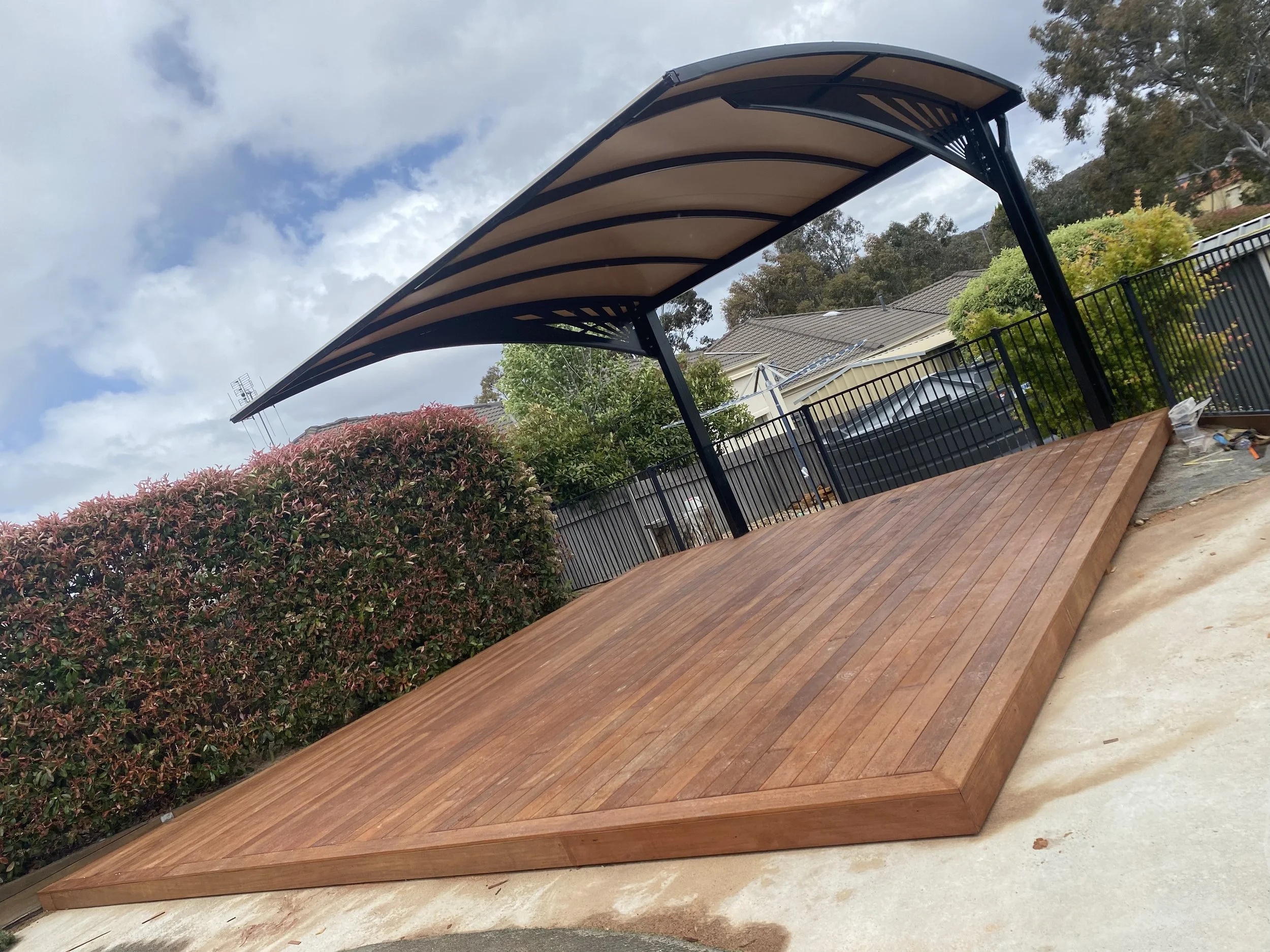 Wooden stage with curved roof in an outdoor setting, surrounded by bushes and fence.