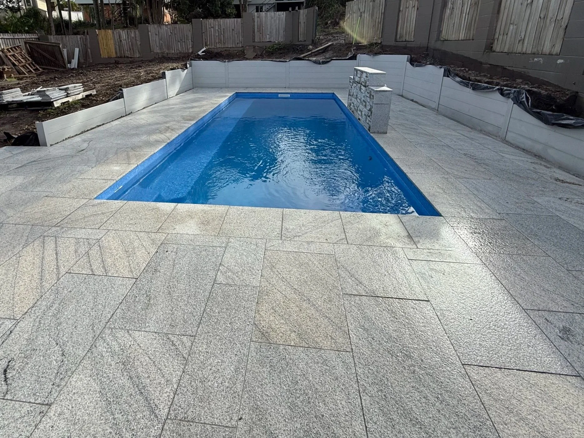 A newly constructed backyard swimming pool with a blue liner, surrounded by beige textured paving stones, with a half-built wall on one side and a wooden fence in the background.