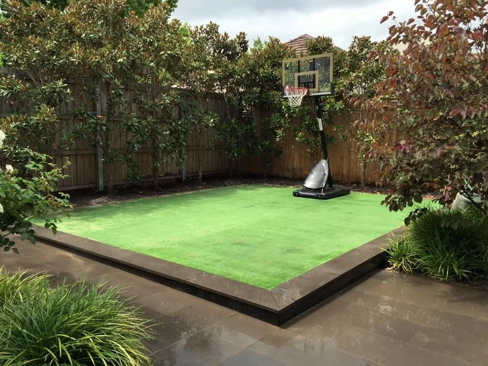 Backyard with a small basketball court and basketball hoop, surrounded by plants and a wooden fence.
