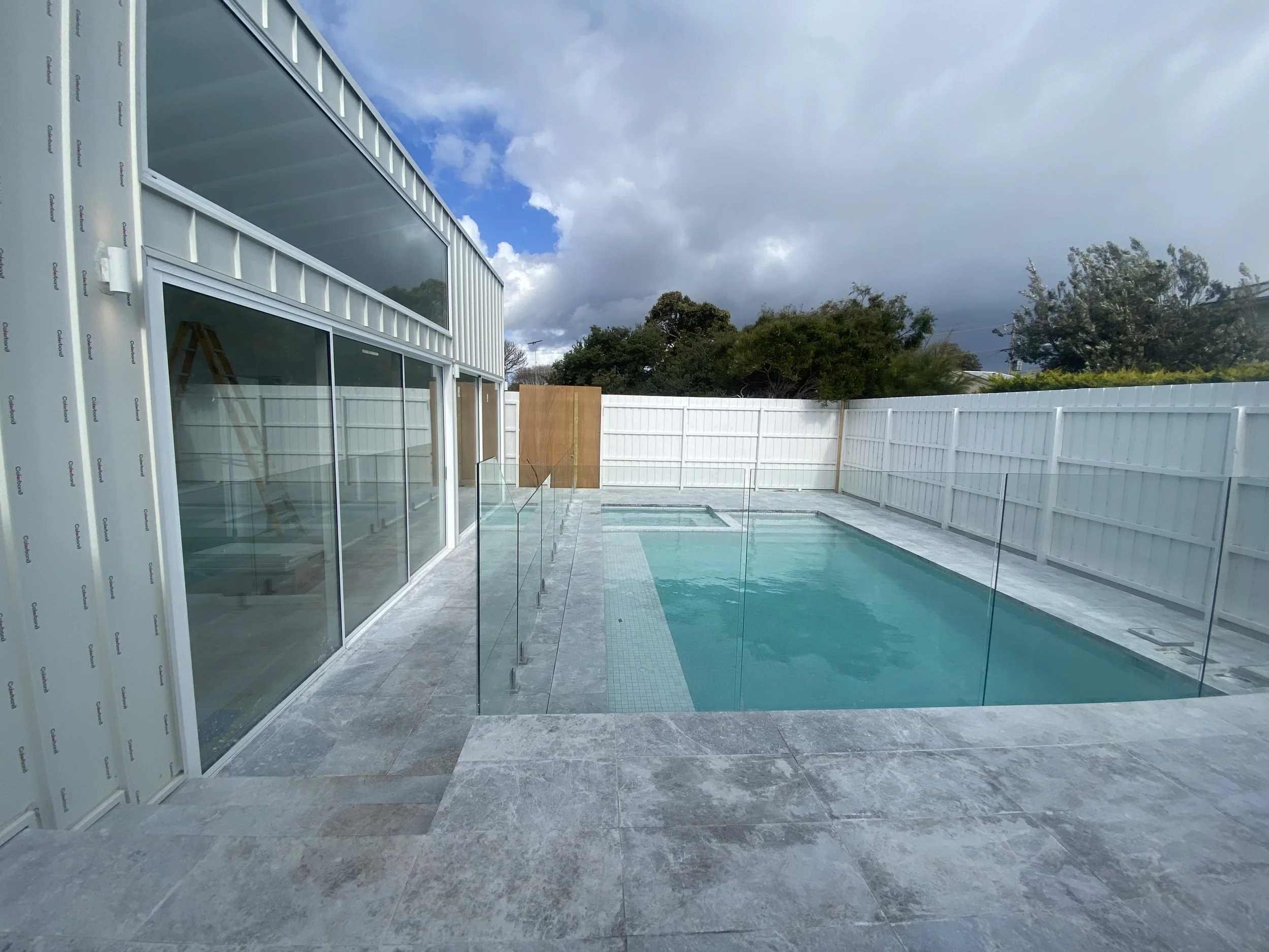 A backyard pool area with a fenced glass barrier, adjacent to a modern house with large glass sliding doors, and an unfinished building with exposed framing. The sky is partly cloudy with patches of blue, and trees are visible in the background.