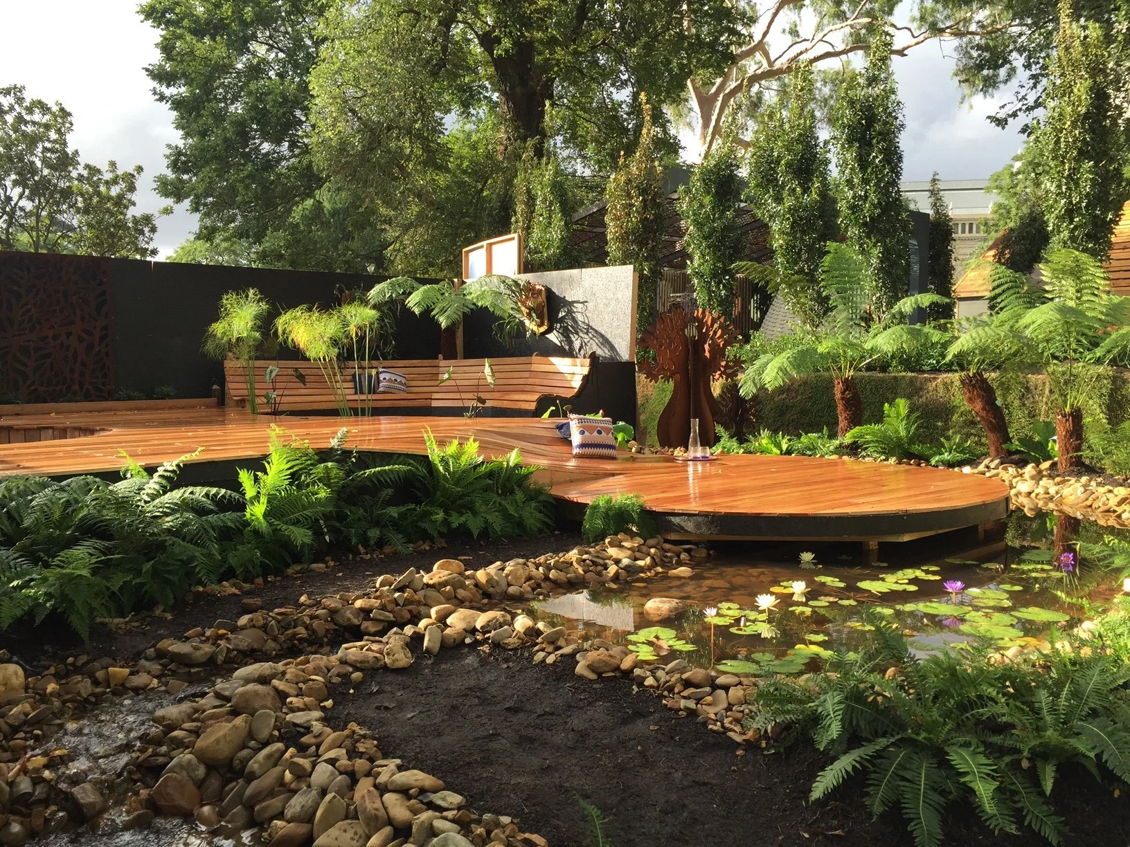 A landscaped garden with a curved wooden deck, lush green plants, small trees, water lilies on a pond, decorative pillows on the deck, and large trees in the background.