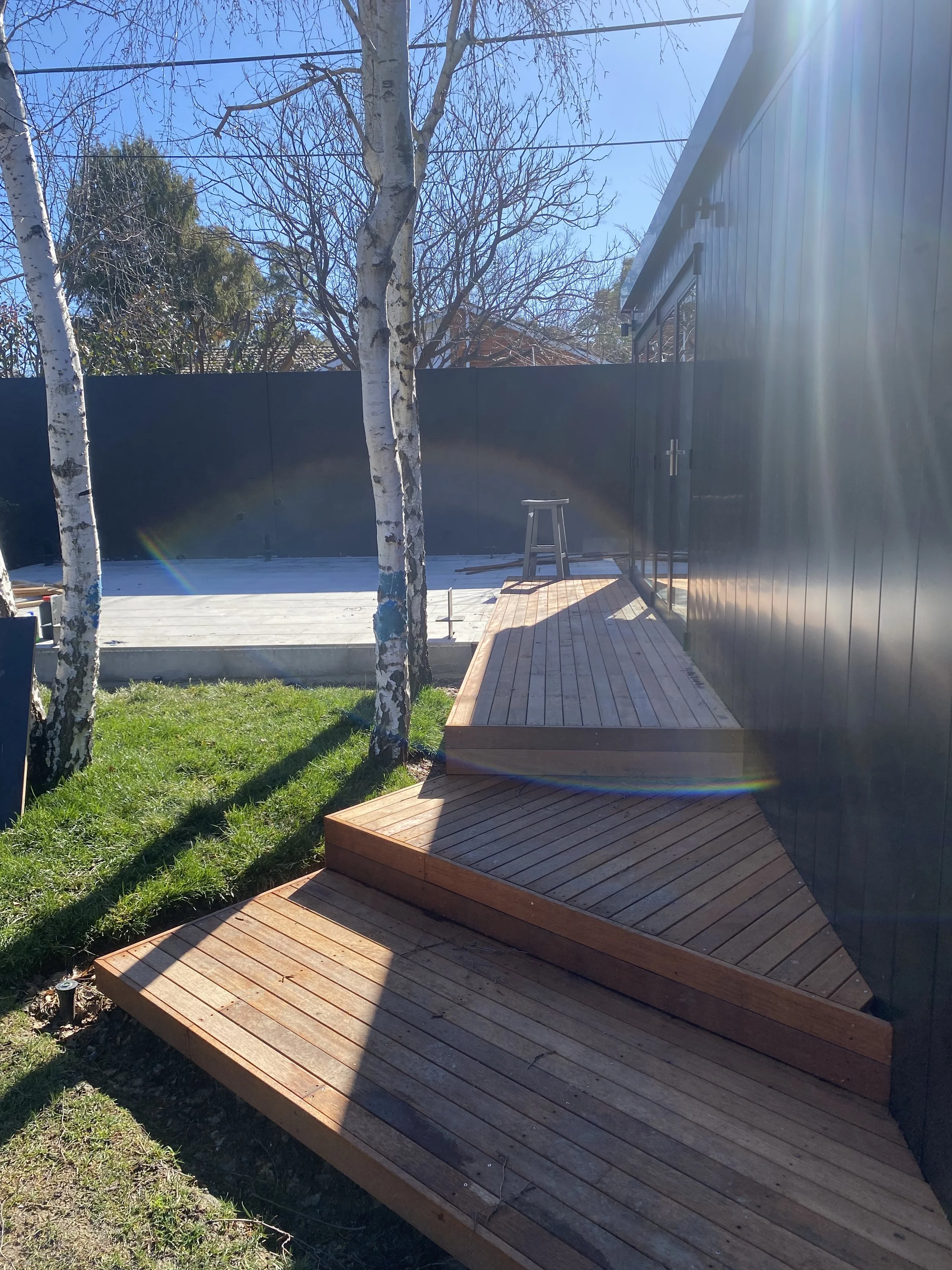 New wooden deck with steps next to a black siding wall, with a small stool on the deck, trees in the background, clear blue sky, and sunlight creating lens flares.
