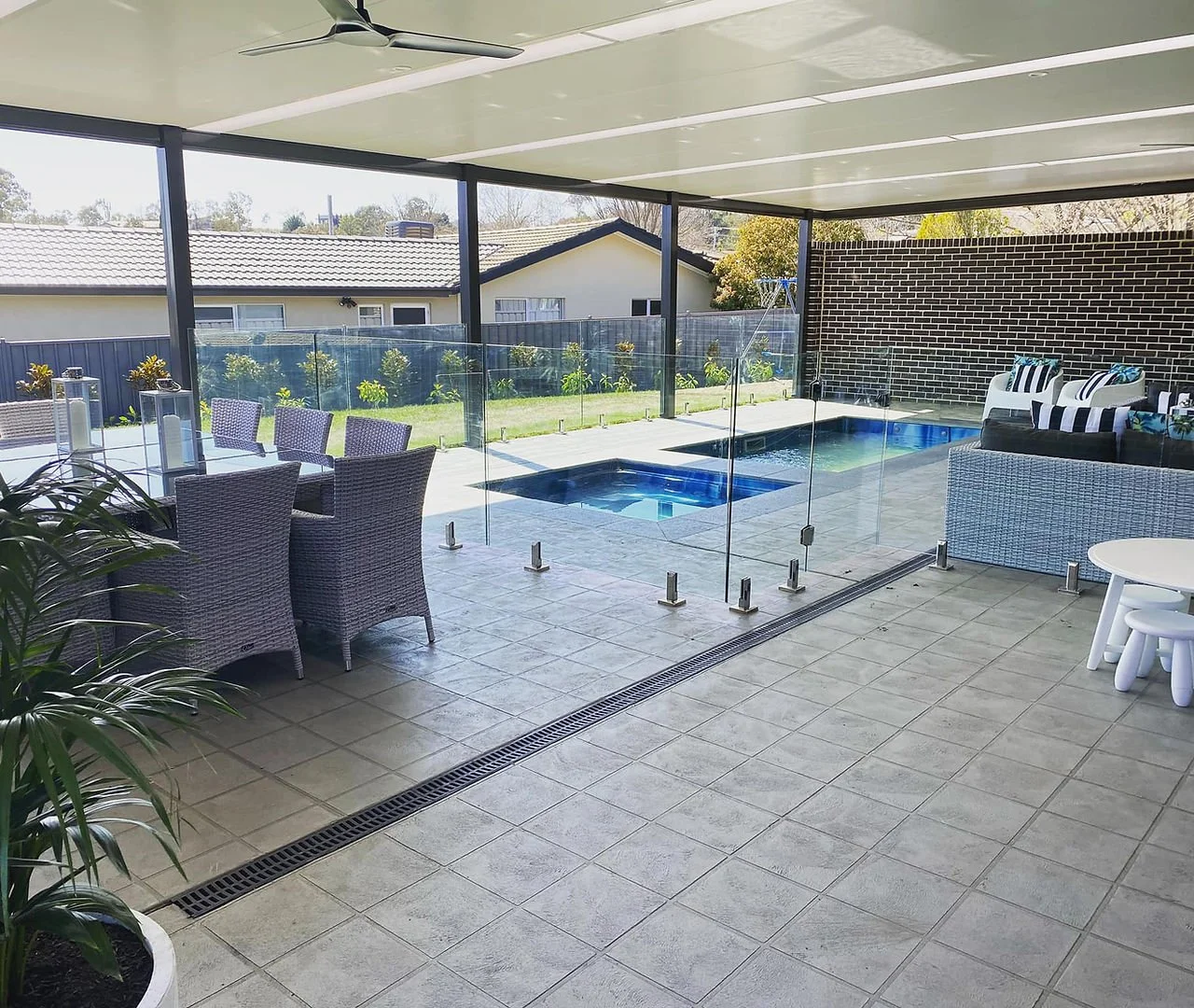 Covered outdoor patio with tiled floor, glass fence, a small pool, outdoor seating including wicker chairs, a wicker lounge, and a round white table with stools, overlooking a grassy yard and neighboring houses.