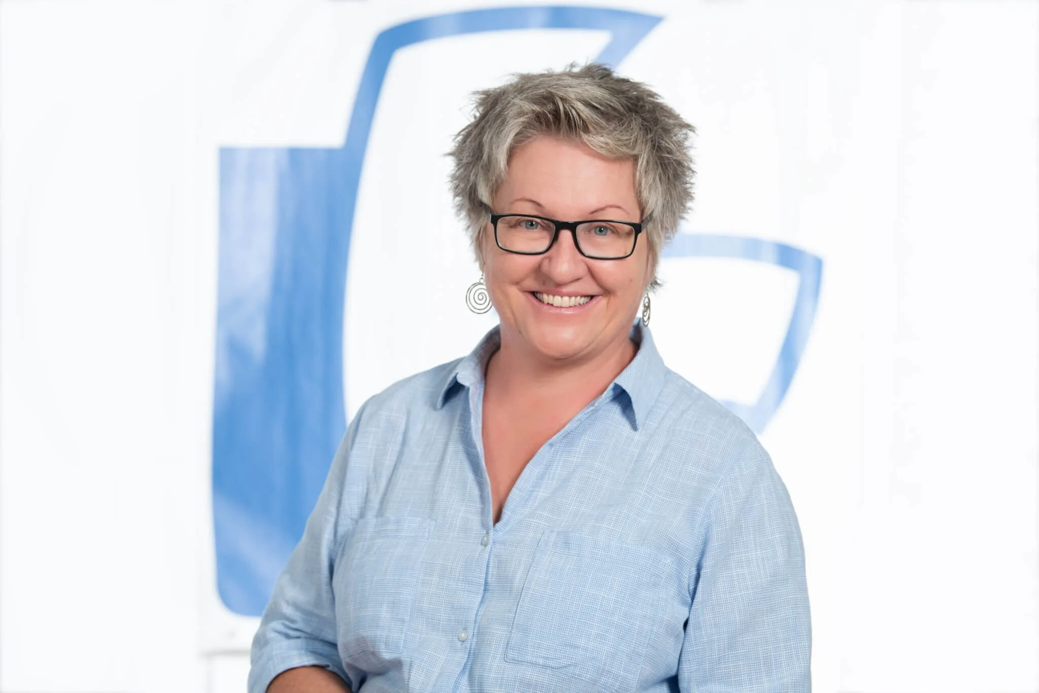 Lächelnde Frau mit Kurzhaarfrisur, Brille und blauer Hemdbluse vor einem weißen Hintergrund mit blauer Logo-Projektionsfolie.