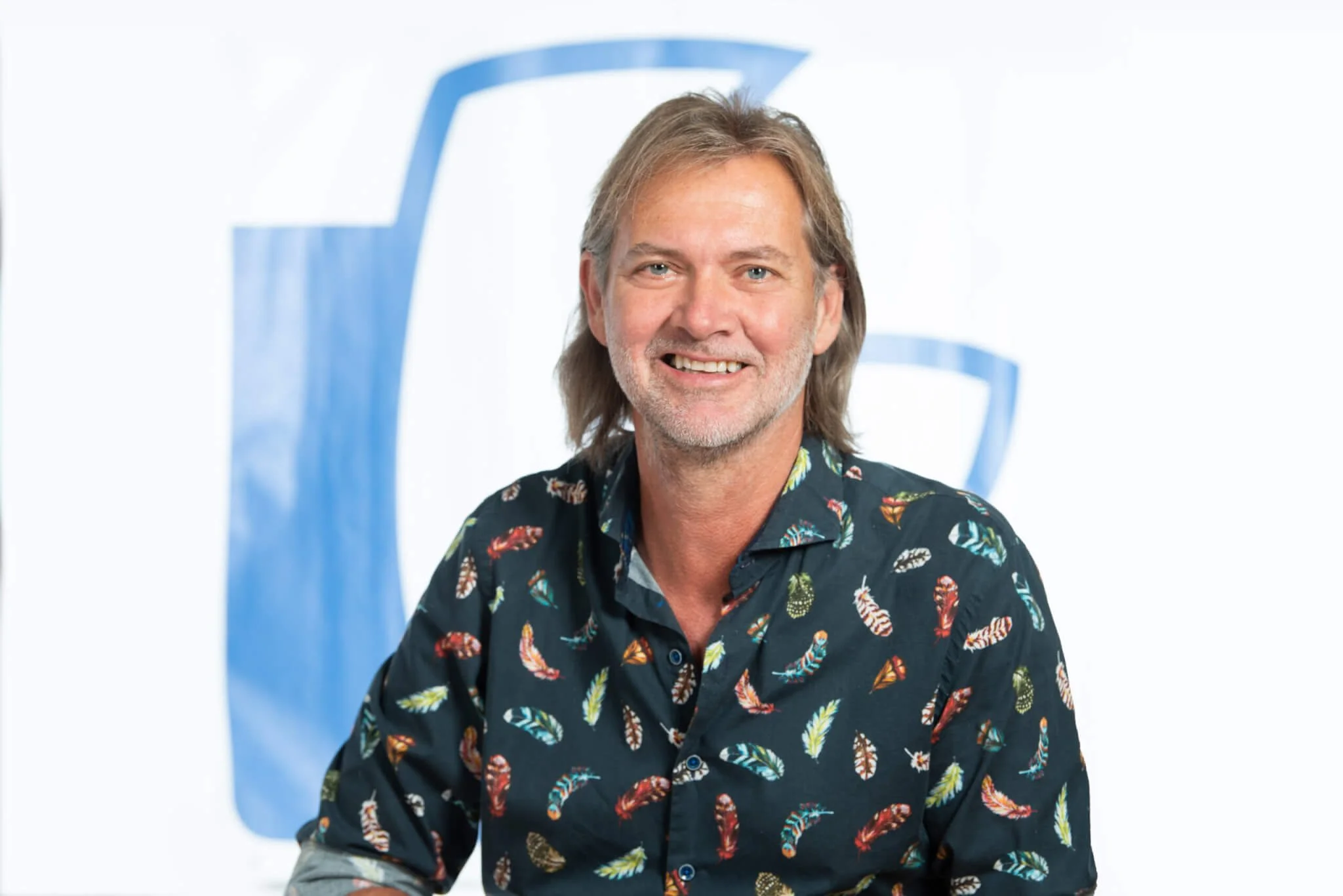 Ein lächelnder Mann mit länglichem, leicht grauem Haar trägt ein Hemd mit bunten Federn auf dunklem Hintergrund. Im Hintergrund ist ein großes, blaues Logo auf weißem Grund.