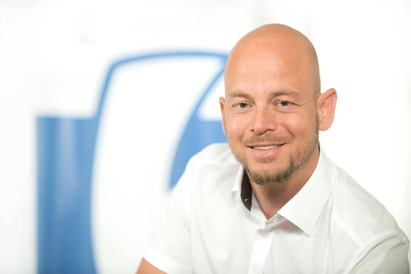 Porträt eines lächelnden Mannes mit Glatze, weißem Hemd, vor einem Hintergrund mit blauen und weißen Elementen.