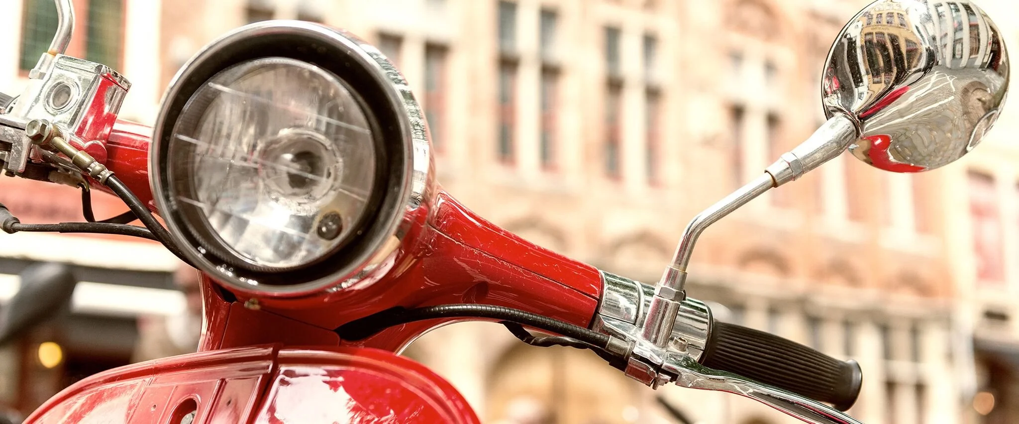 Nahaufnahme des Vorderrads und Lenkers eines roten Motorrollers mit Spiegel vor einer städtischen Hintergrund mit alten Backsteingebäuden.