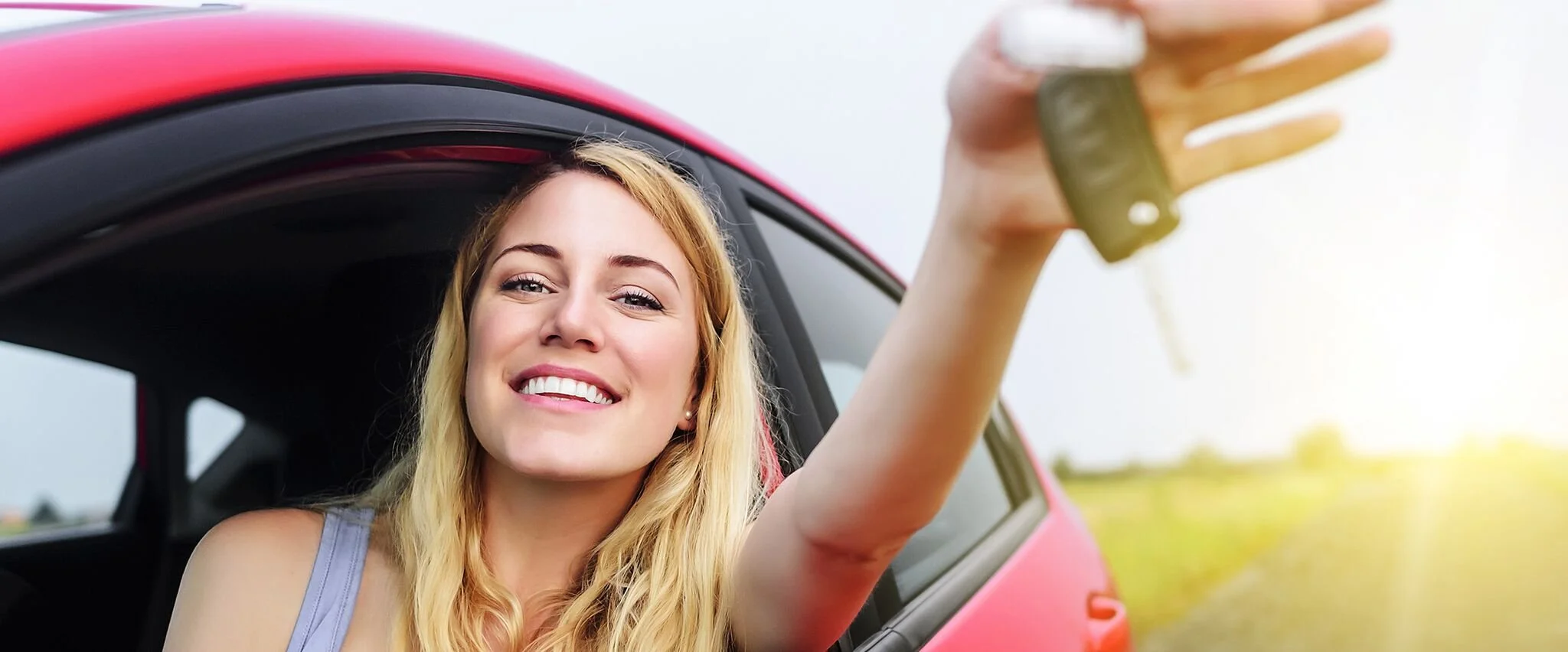 Eine junge Frau, die lachend aus dem Autofenster eines roten Autos schaut, während sie einen Autoschlüssel hochhält.