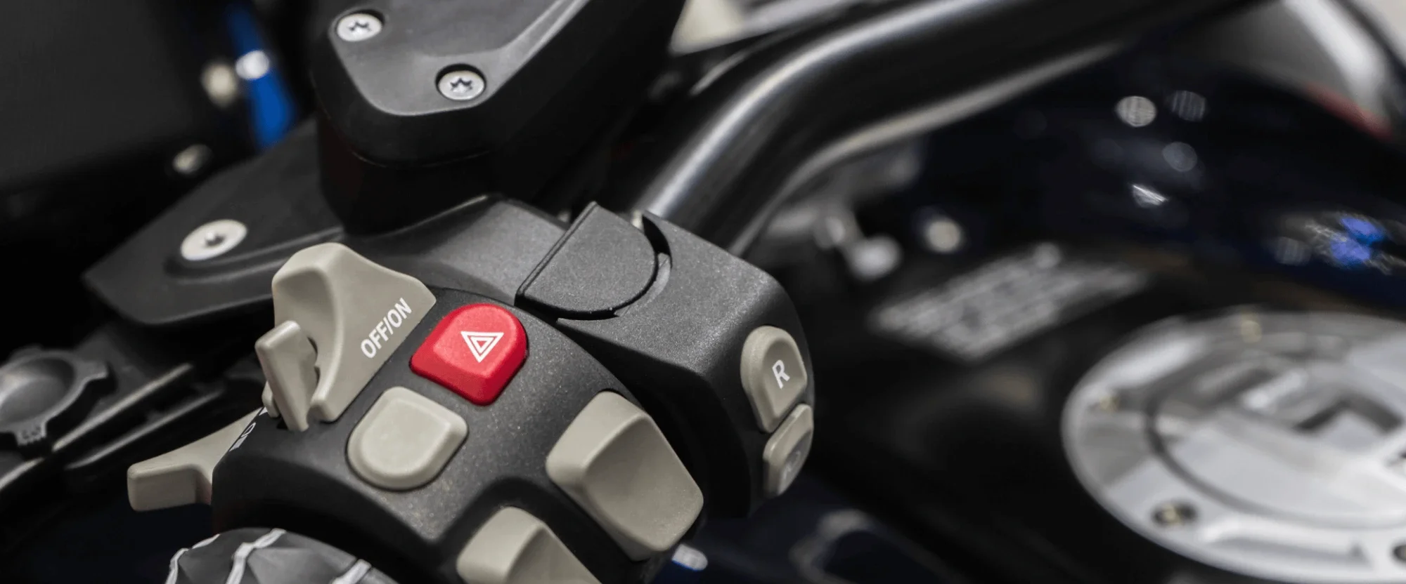 Nahaufnahme eines Motorradlenkers mit mehreren Bedienelementen, einschließlich des roten Warnblinklicht-Buttons, eines Off-Buttons und eines R-Buttons, mit sichtbaren metallischen Teilen im Hintergrund.