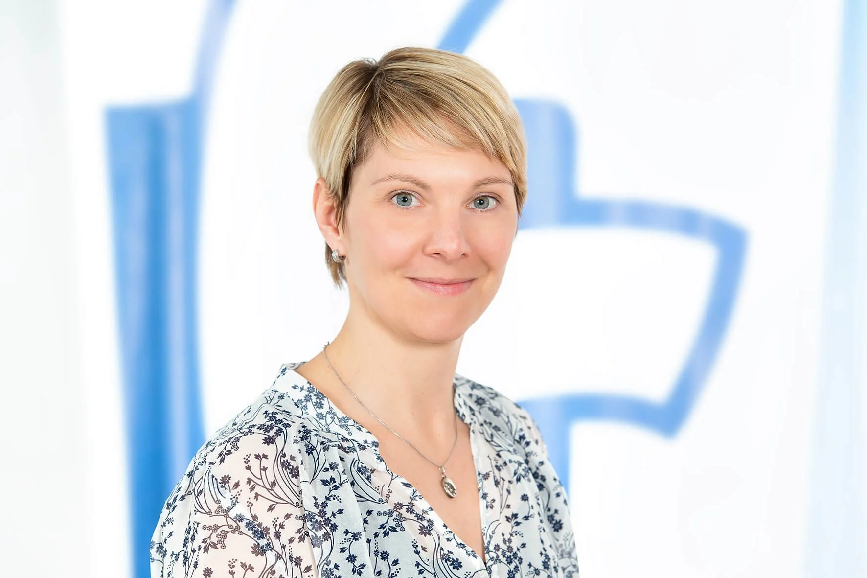 Porträt einer Frau mit kurzen, blonden Haaren vor einem weißen Hintergrund mit blauer Grafik.
