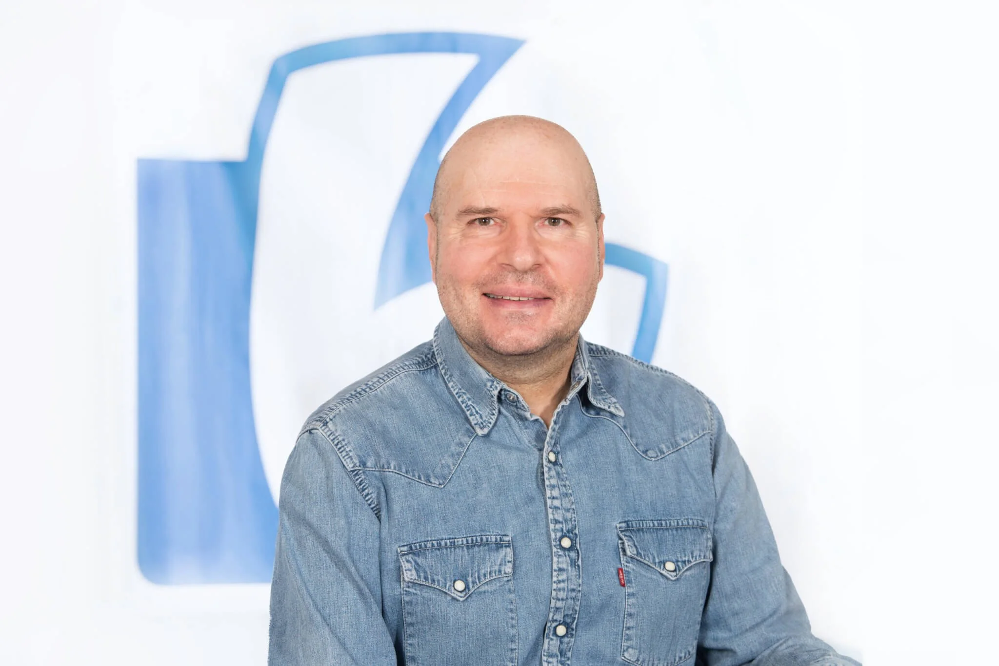 Mann mit heller Haut, blassem Gesicht und kurz geschnittenem Haar, trägt ein denim Hemd, sitzt vor einem weißen Hintergrund mit einem blauen abstrakten Logo.