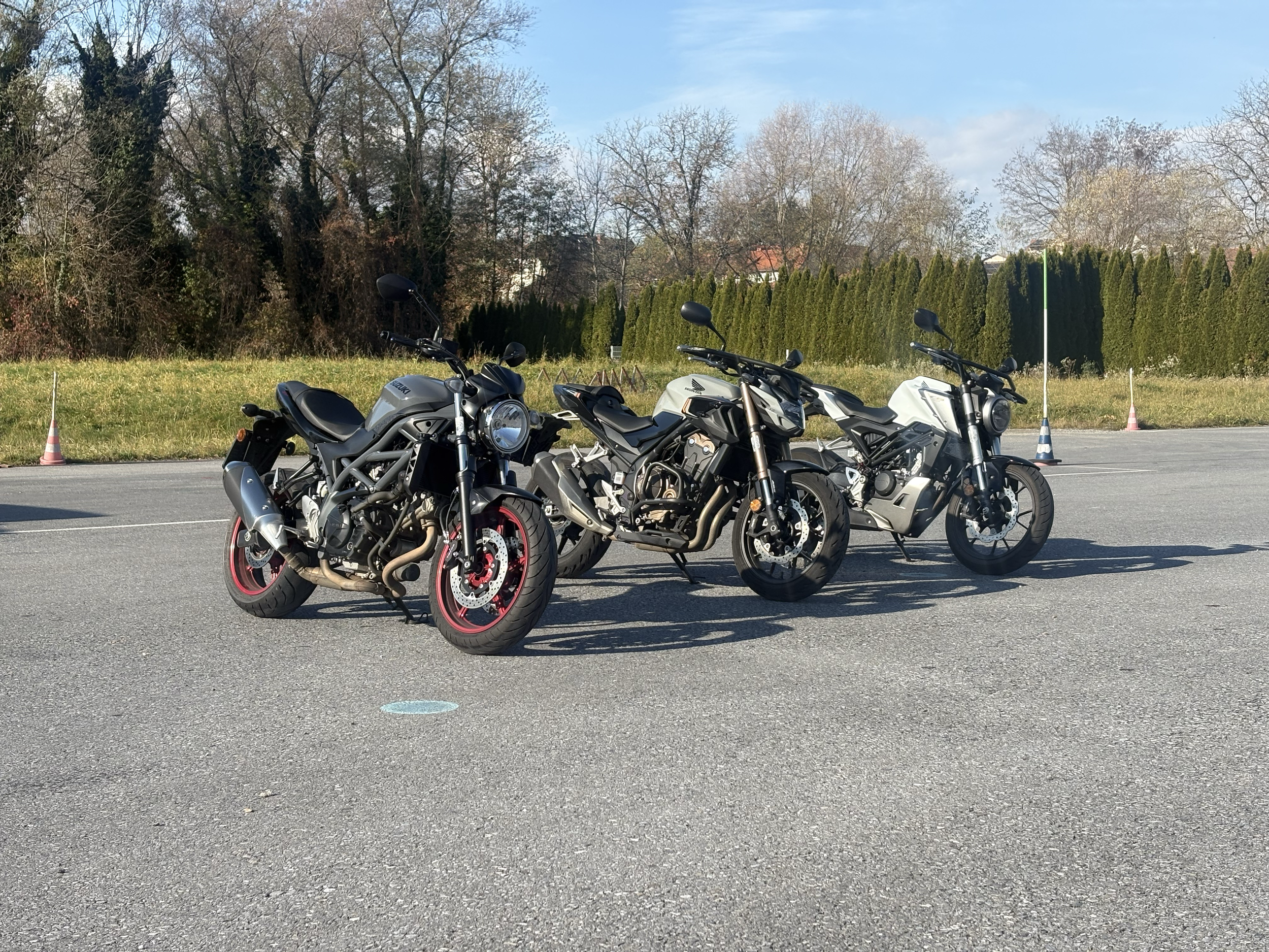 Drei Motorräder stehen nebeneinander auf einem asphaltierten Parkplatz, hinter ihnen ein Grünstreifen und eine Hecke, und im Hintergrund Bäume unter einem blauen Himmel.