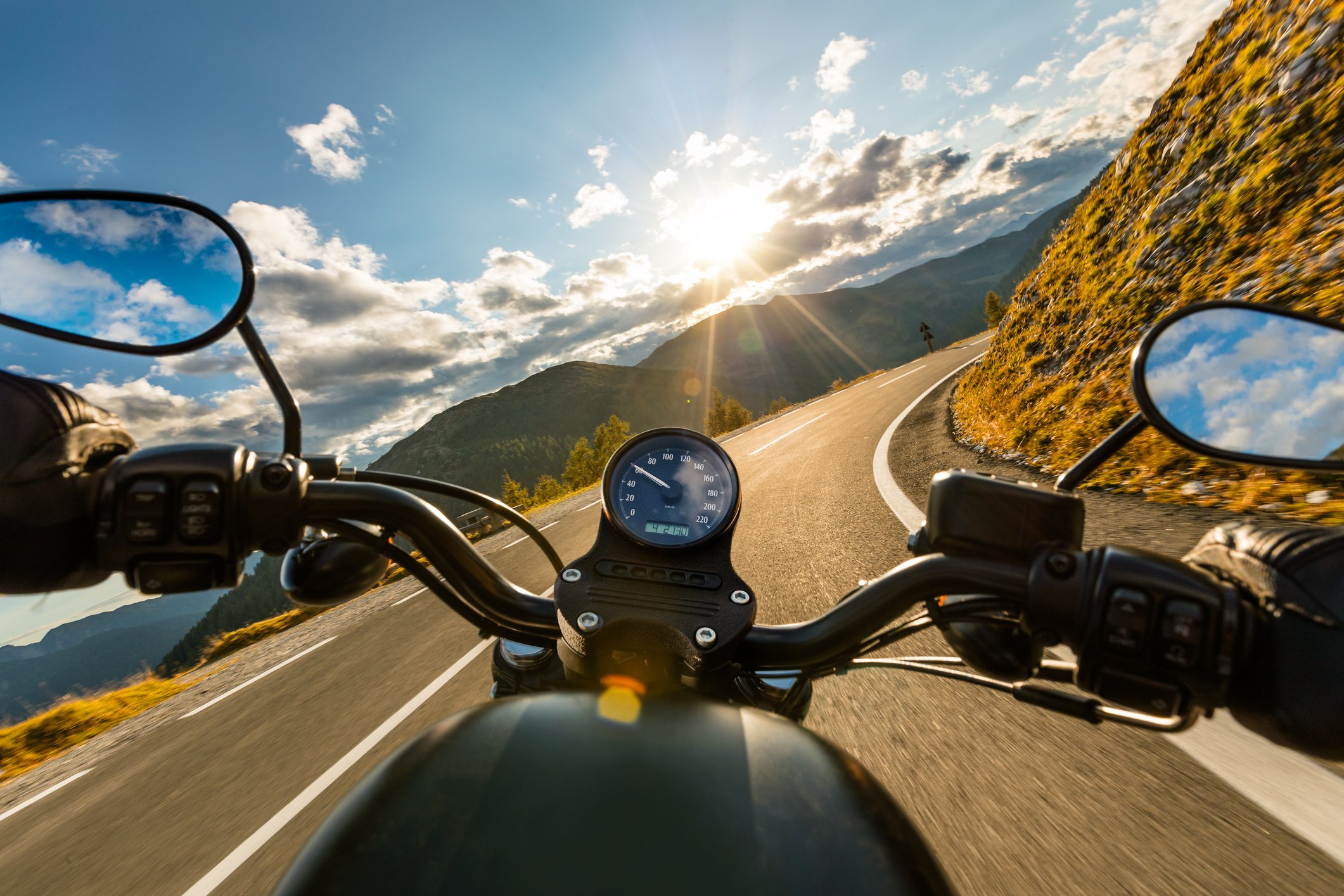 Motorradfahrer fährt auf kurviger Straße in den Bergen bei Sonnenuntergang, Blick vom Lenker auf Tacho und Spiegel, umgeben von Sonnenlicht und Wolken