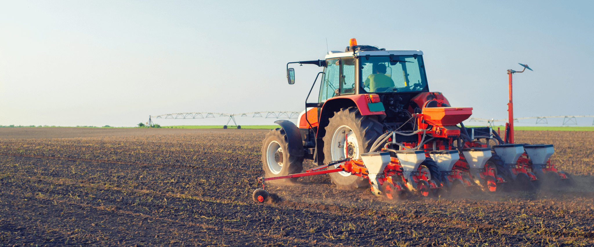 Ein roter Traktor arbeitet auf einem Acker, der Boden wird gepflügt oder vorbereitet für die Aussaat, im Hintergrund sind Bewässerungssysteme sichtbar.