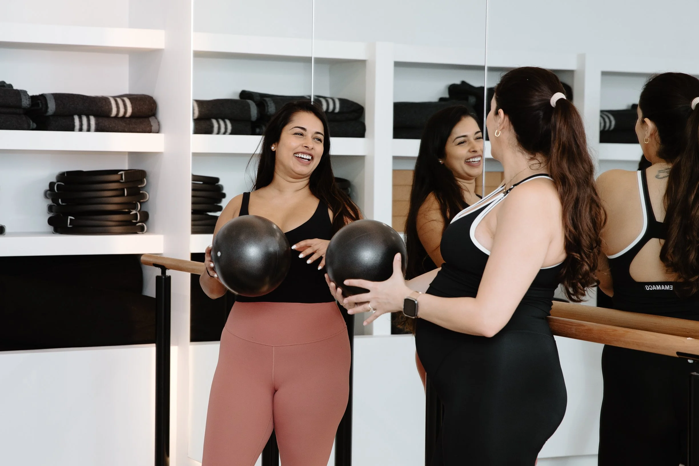 Two women in workout attire holding black exercise balls and smiling at each other in a fitness studio with Shelves of towels and belts behind them.