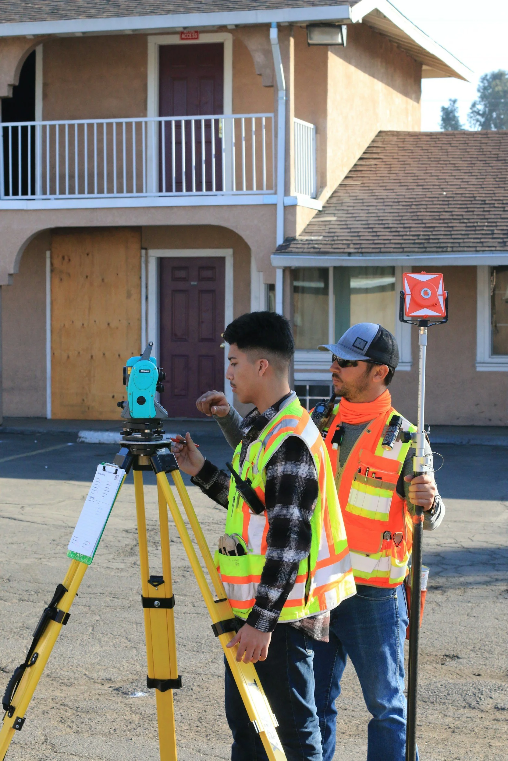Two construction workers in safety vests using surveying equipment outside a building with boarded-up windows. Clean-Up
