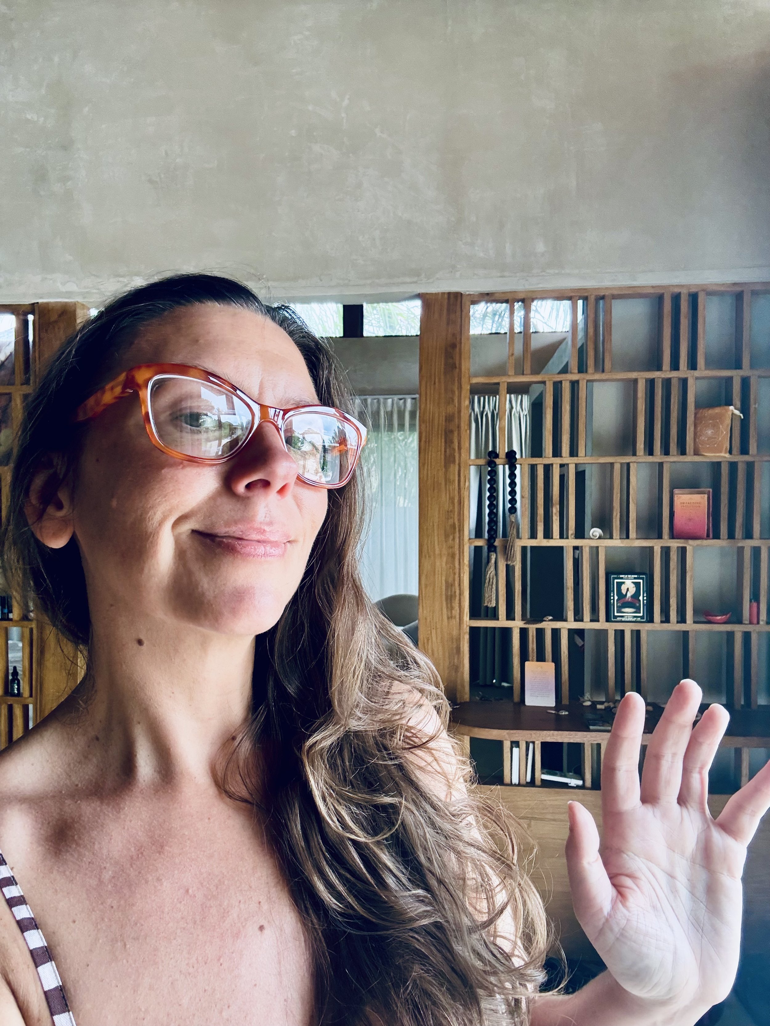 A woman with long wavy hair wearing large tortoise-shell glasses, a striped tank top, and light makeup, smiling slightly and raising her right hand with fingers slightly spread, in front of a wooden partition with decorative items and books inside a modern styled room.