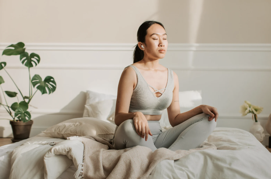 A woman practicing meditation in a bedroom, sitting cross-legged on a bed, with eyes closed and a peaceful expression.
