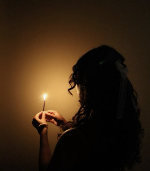 A woman with long curly hair holding a lit sparkler in her hand against a dark background.