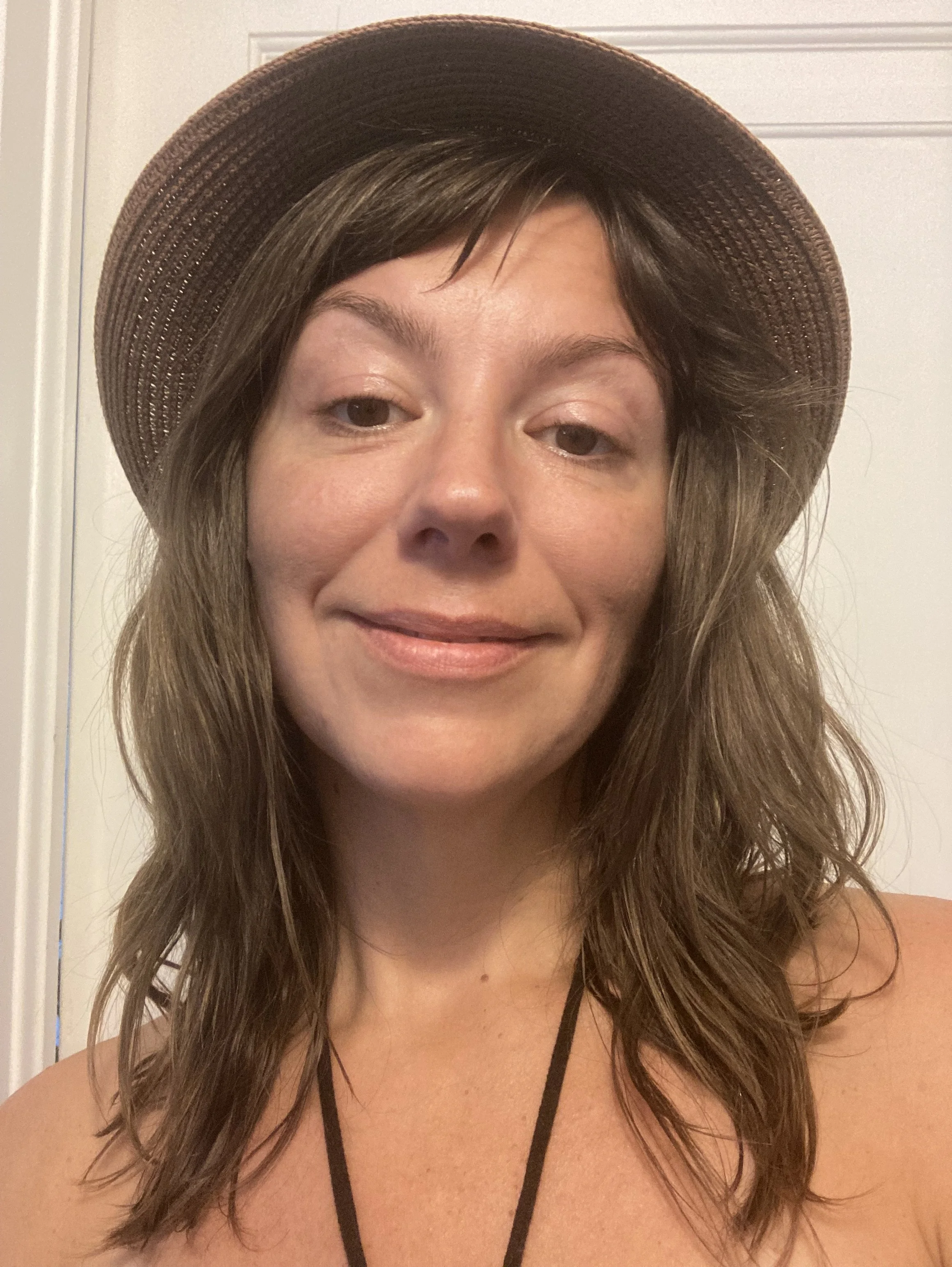 A woman with long wavy hair wearing a large brown straw hat, a black necklace, and a sleeveless top, standing in front of a white door.